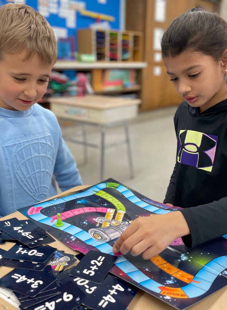 two children playing a board game with math equation cards in a classroom