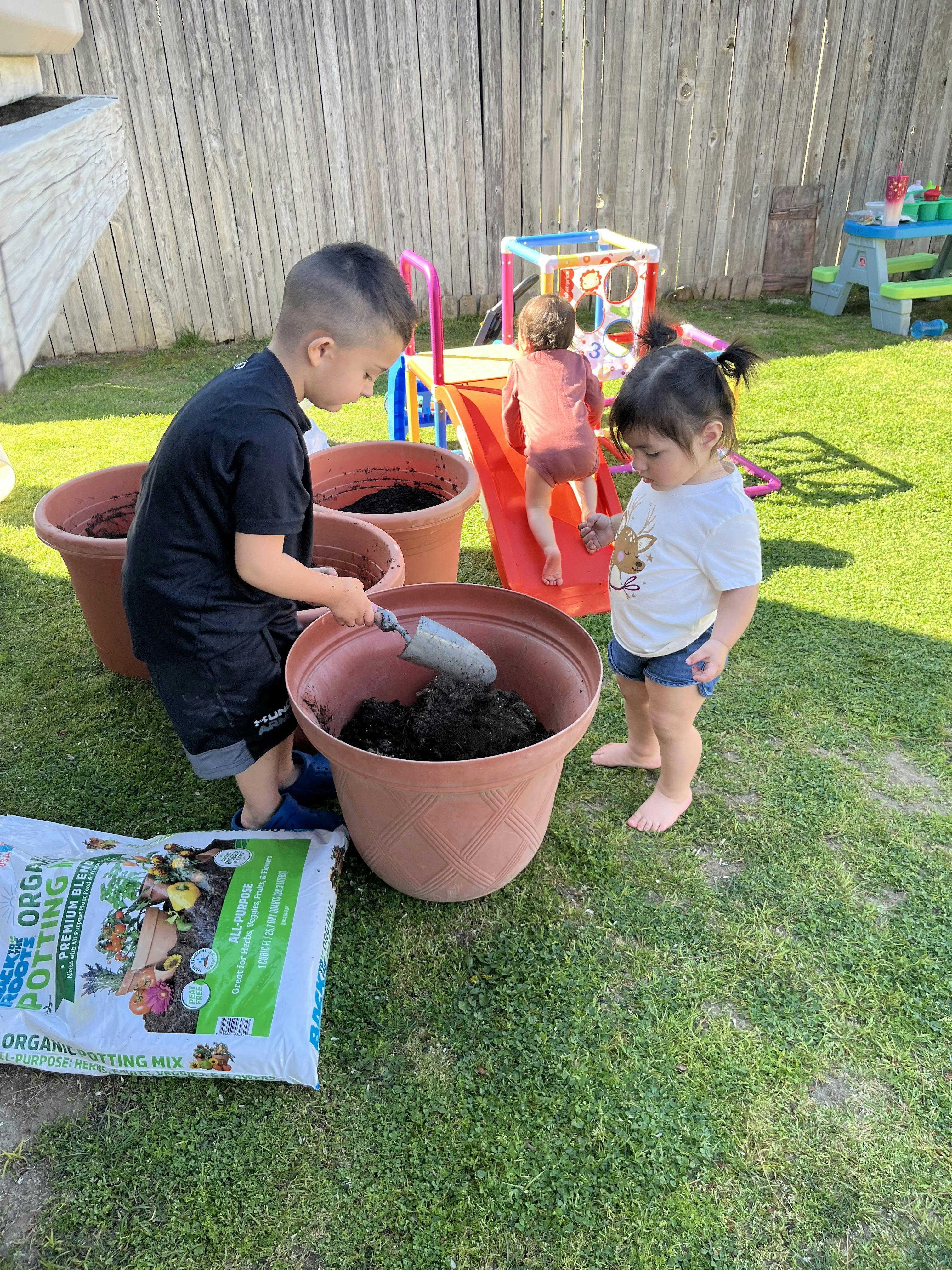 Child using a trowel to put soil into a large pot, another child watching