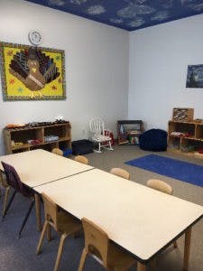 Classroom with tables and chairs, shelves, and a blue rug