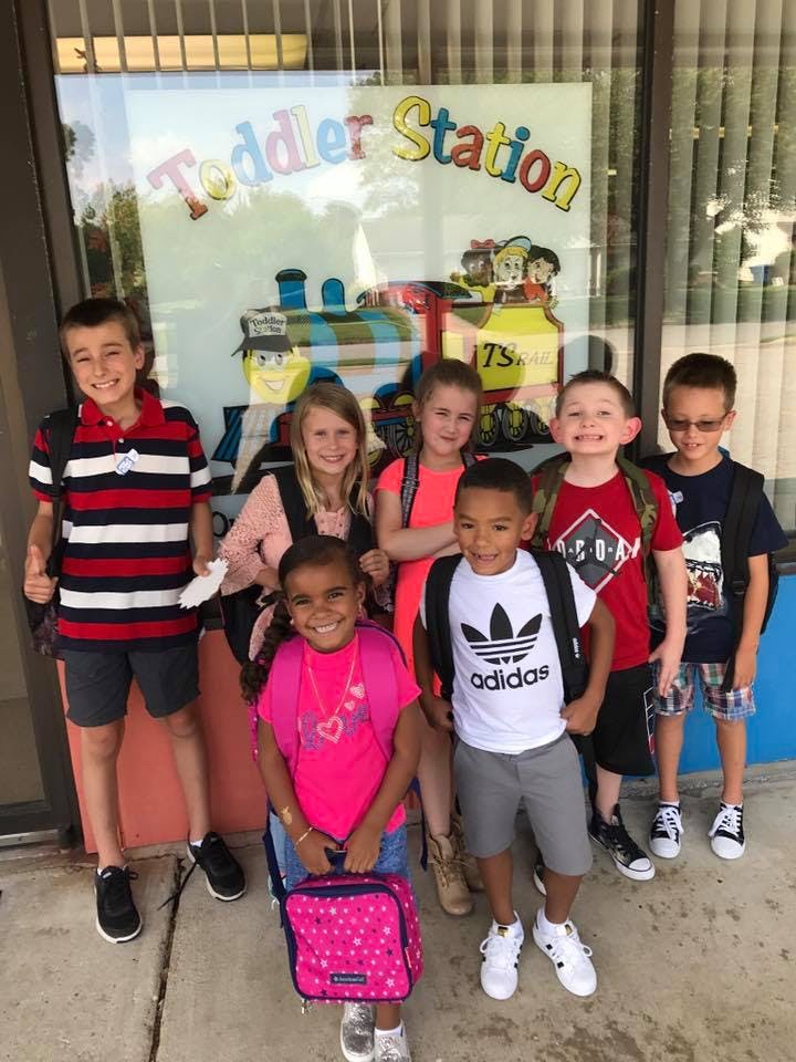 Group of children with backpacks standing in front of a window with "Toddler Station" text