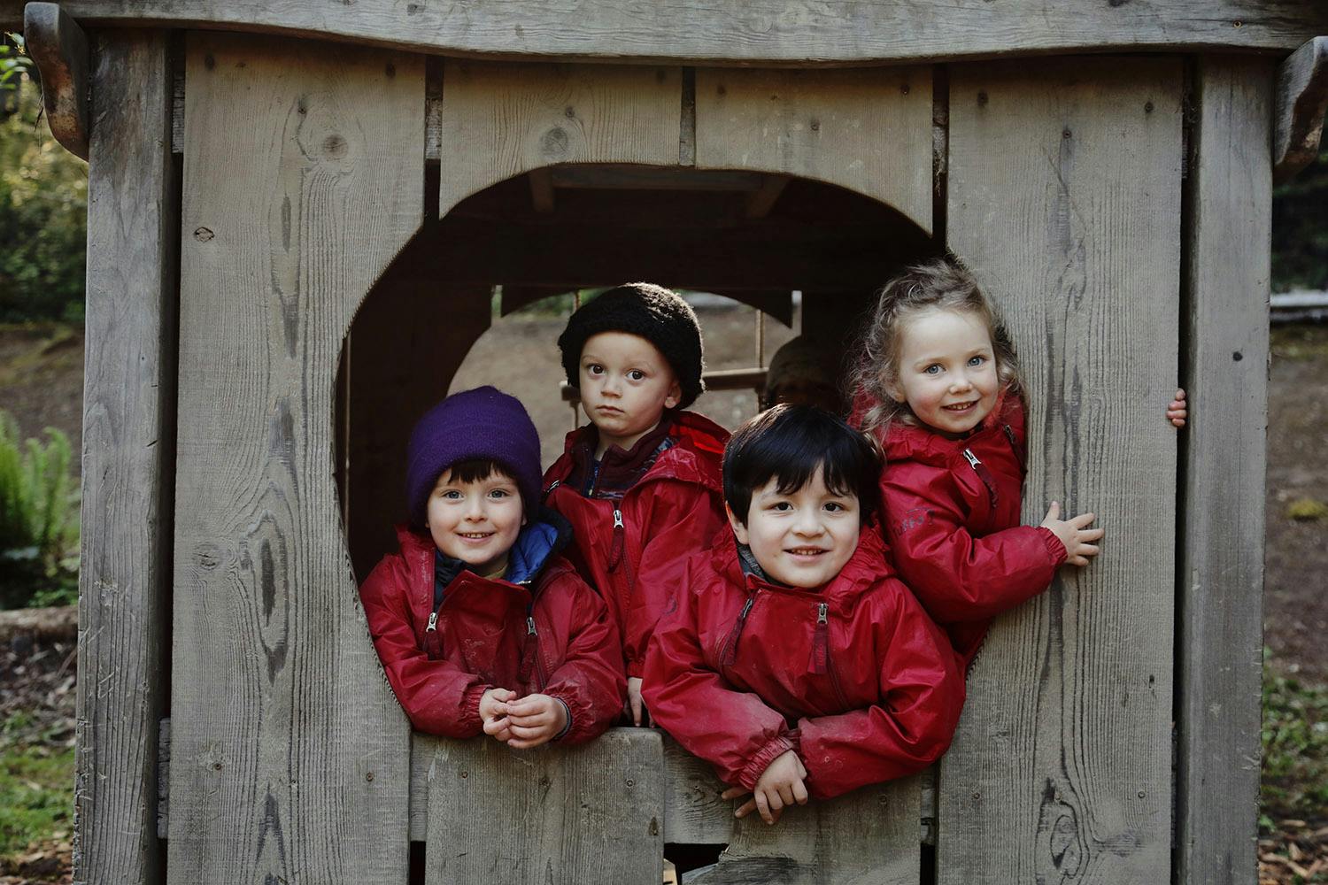 Four children in red jackets looking out from a wooden playhouse