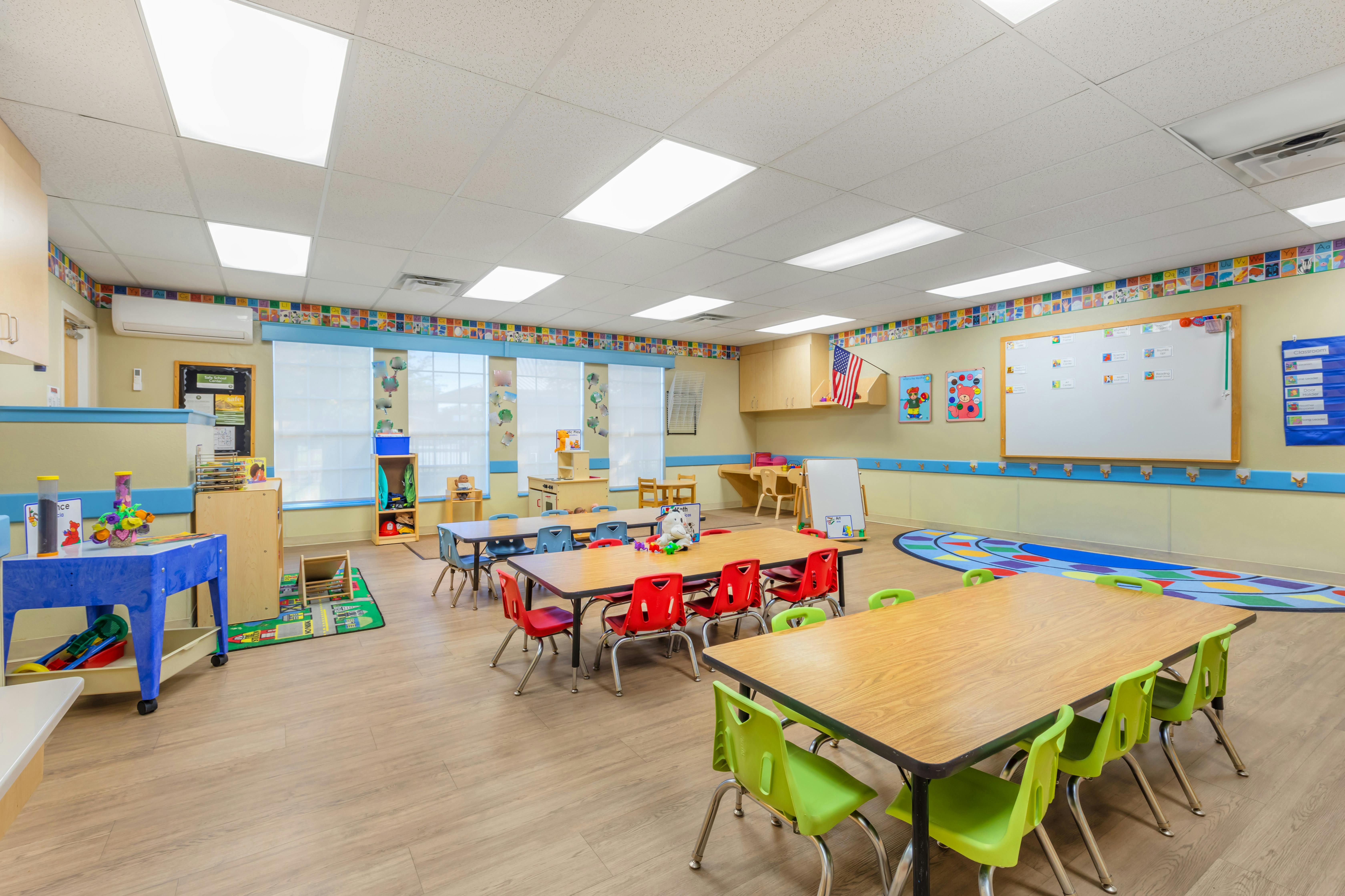 An empty classroom with small tables and chairs, and play mats on the floor