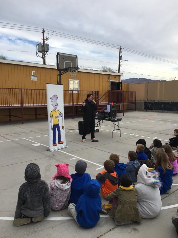 Person speaking to a group of children sitting on the ground outdoors