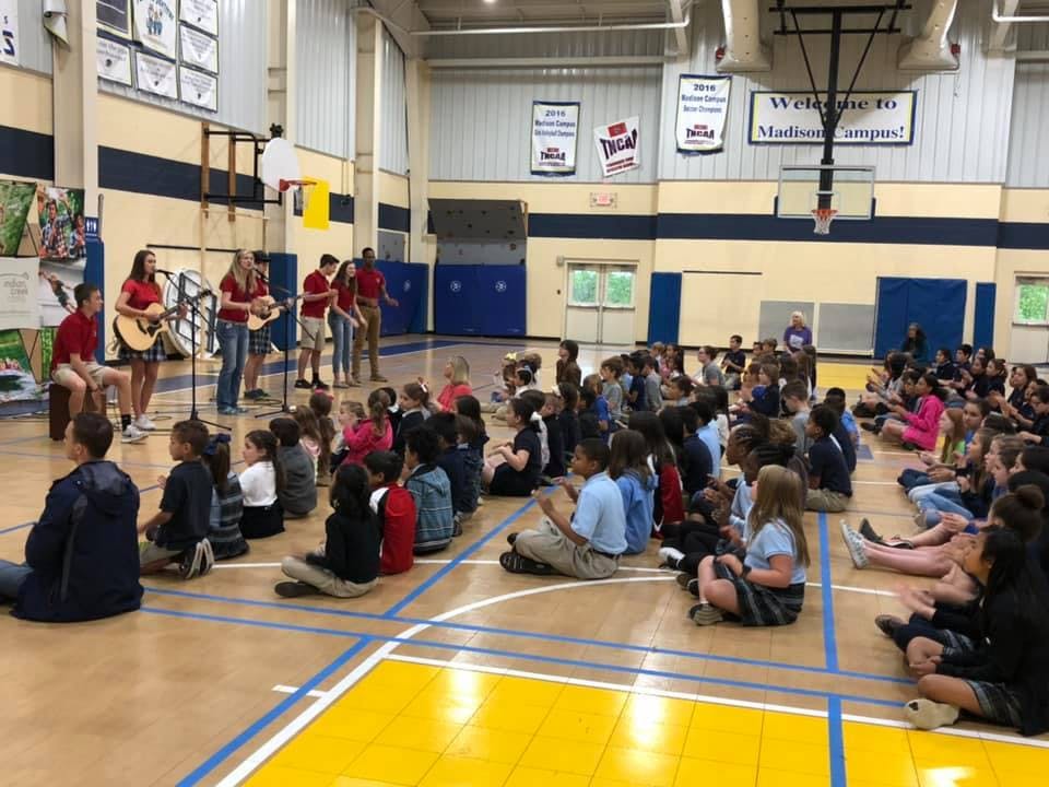 People playing guitars for children sitting on a gymnasium floor