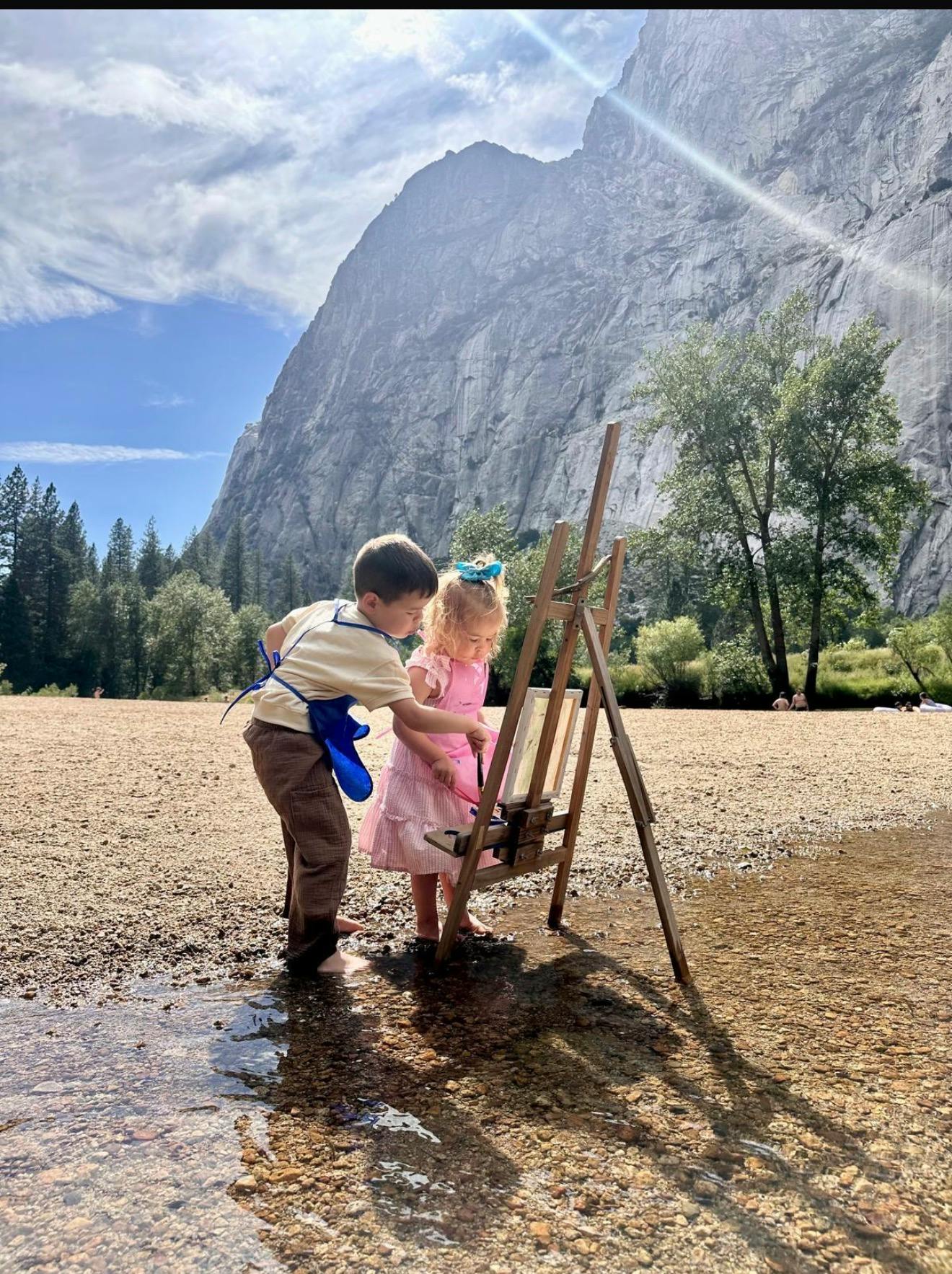 Two children painting on an easel in a rocky riverbed beneath mountains