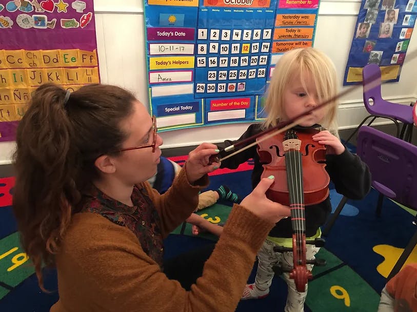 Person helping a child play a violin in a classroom