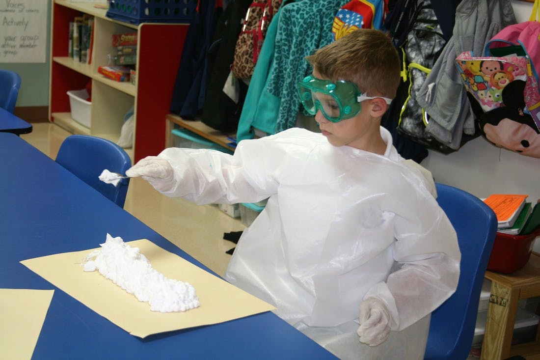 Child in goggles and white lab coat working with white material on paper