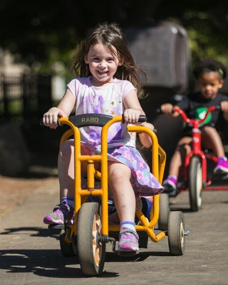 Child riding a yellow tricycle, another child on a red tricycle in background