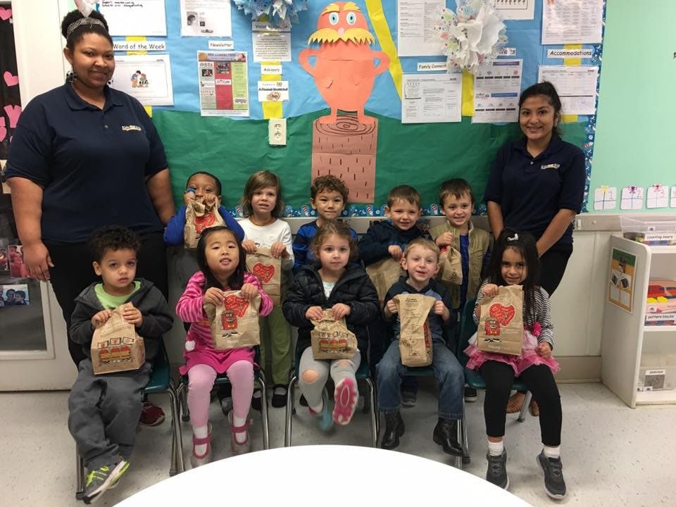 Two people and multiple children holding paper bags in a classroom