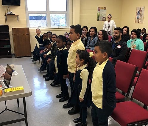 Children standing in rows facing a table with adults seated behind them