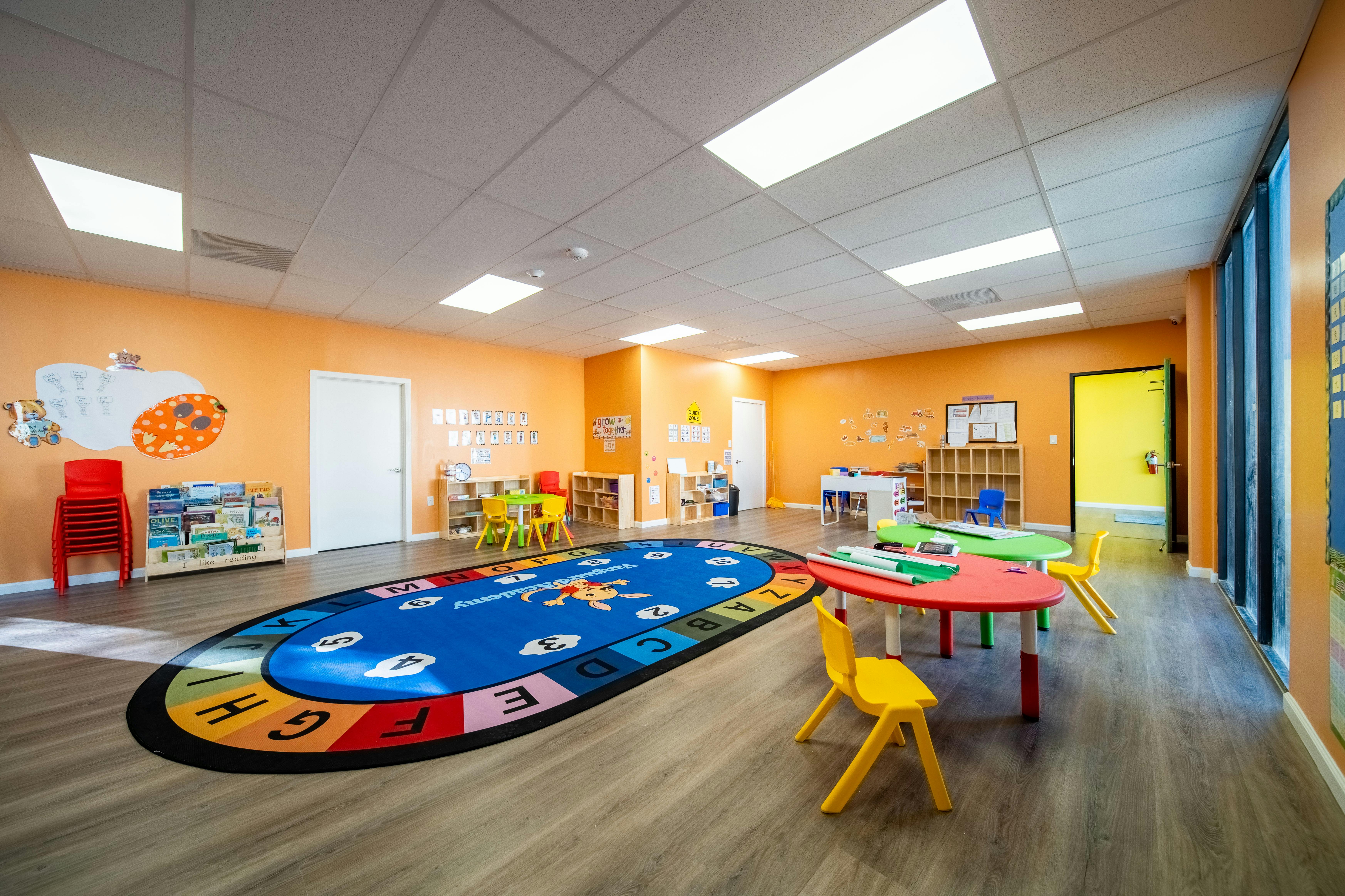 A classroom with small tables and chairs, a large alphabet rug, and bright orange walls