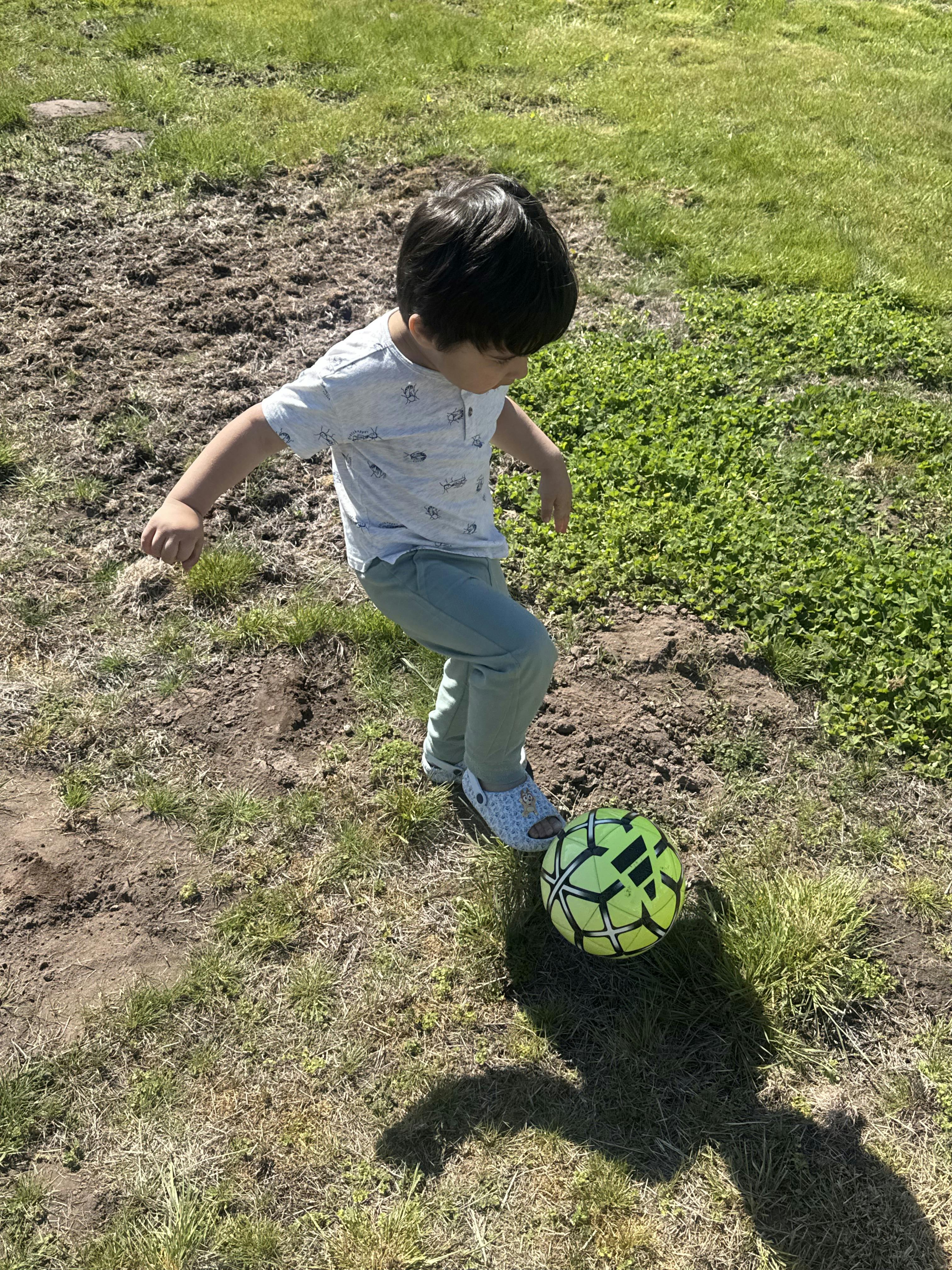 Child about to kick a bright yellow soccer ball on a grassy, muddy patch