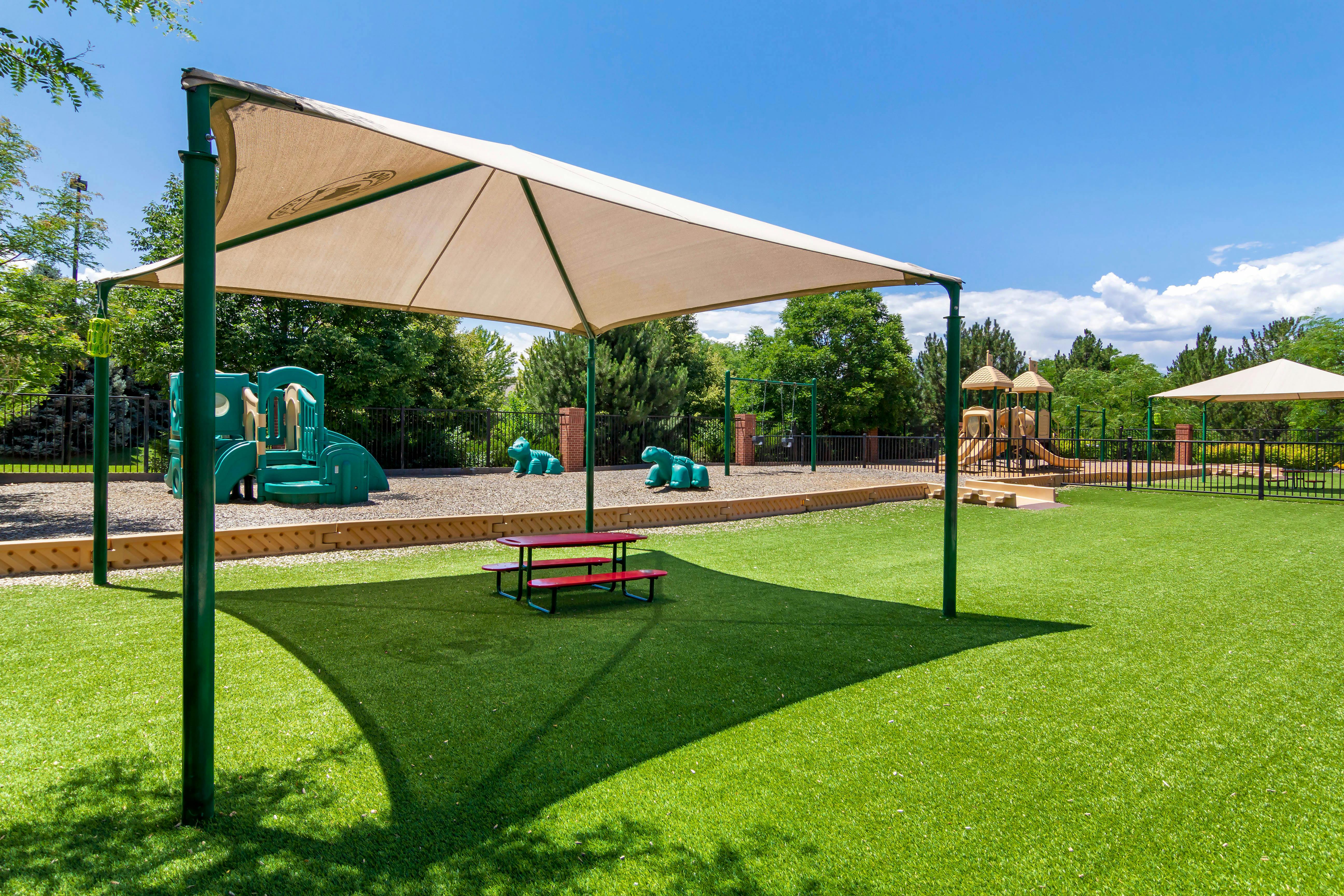 A children's playground with green artificial turf and play structures under a large shade