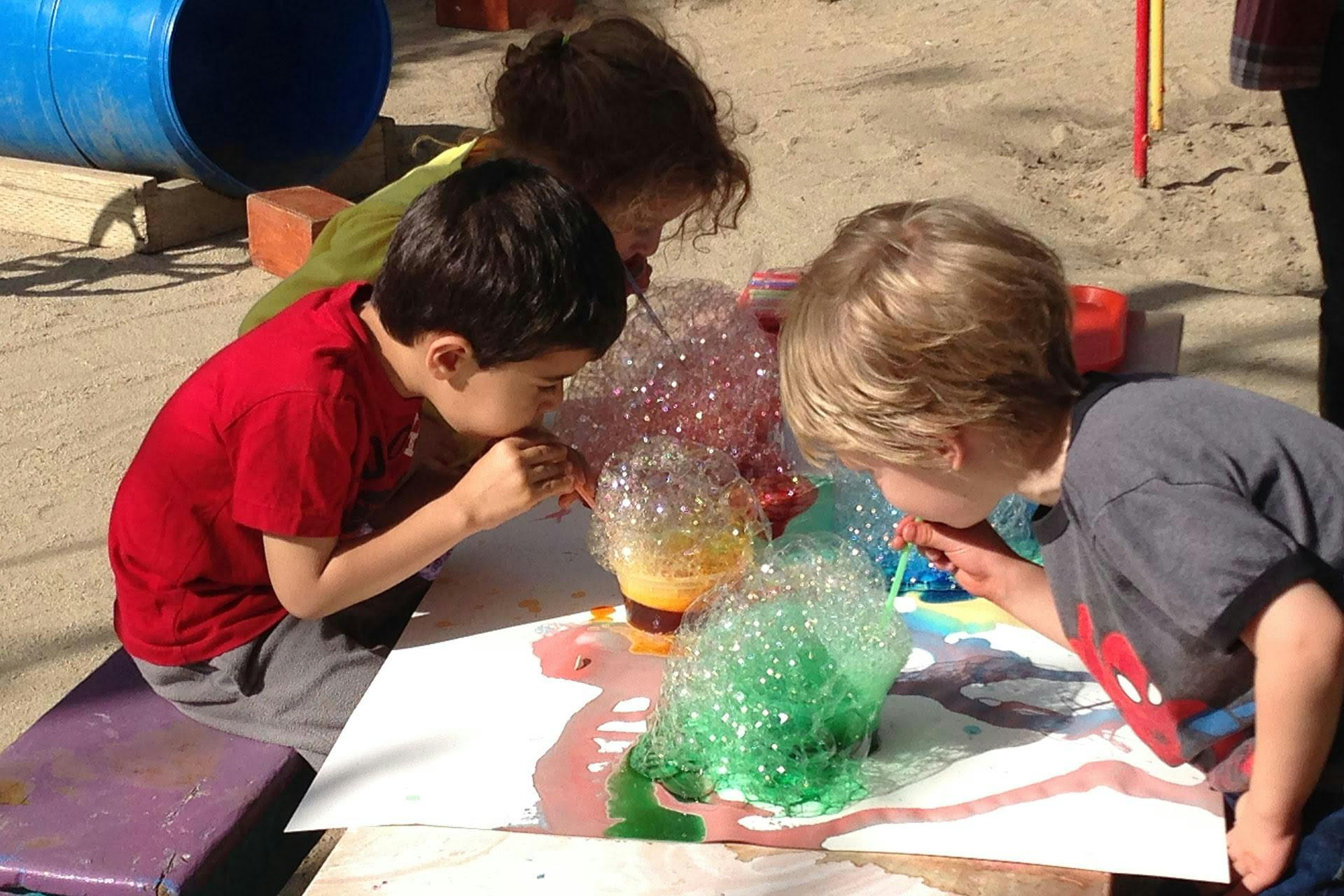 Three children blowing bubbles onto a table with paint on a table