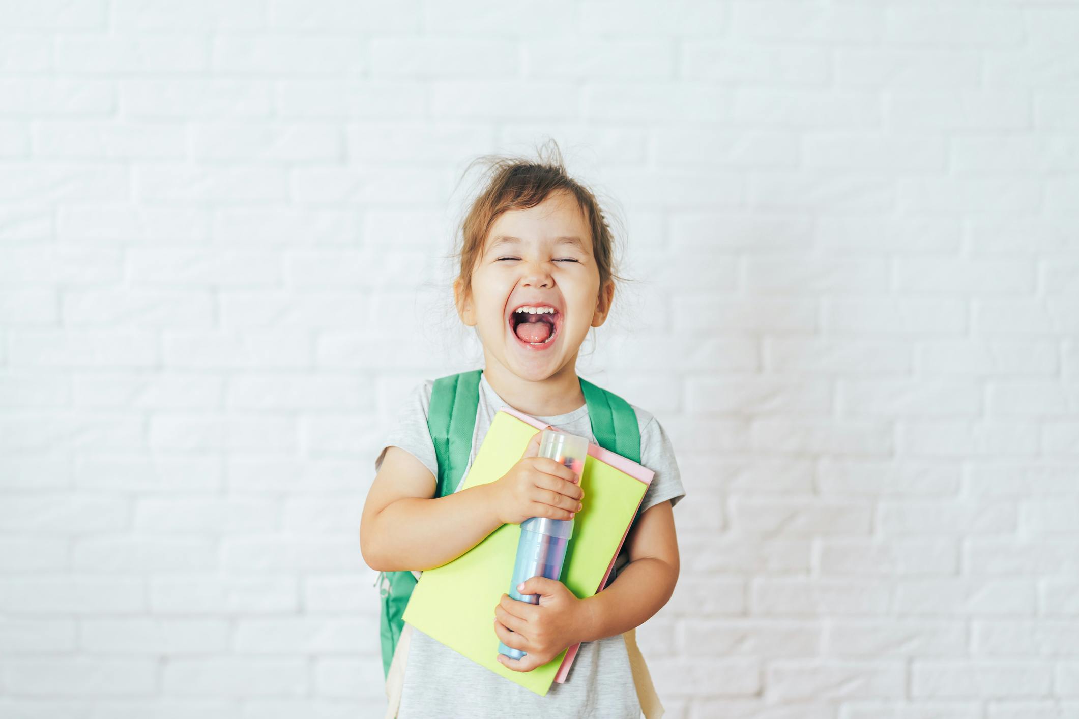 Child wearing a green backpack, holding books and a marker case