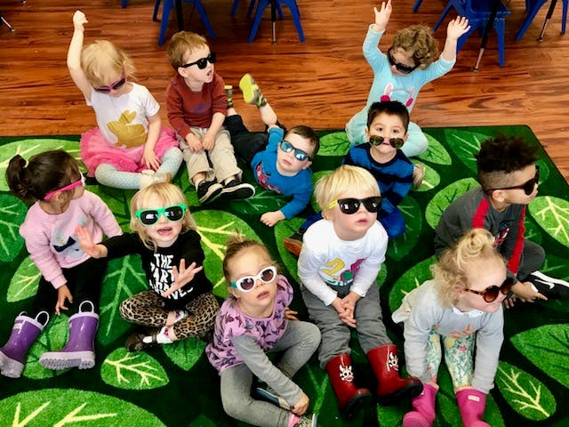 Many children wearing sunglasses sitting and lying on a green rug