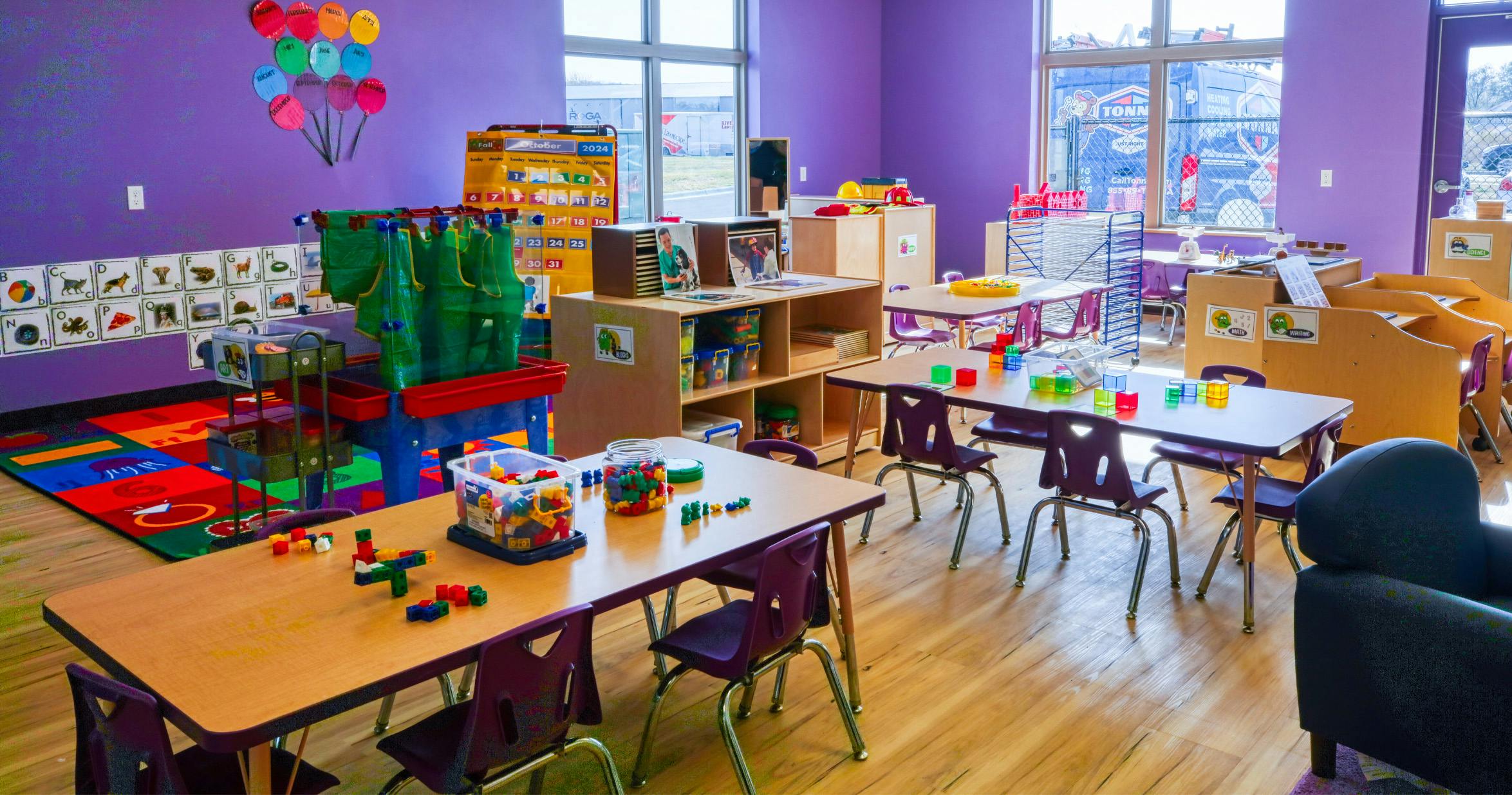 An empty early childhood classroom with multiple tables and chairs, shelves, and learning materials