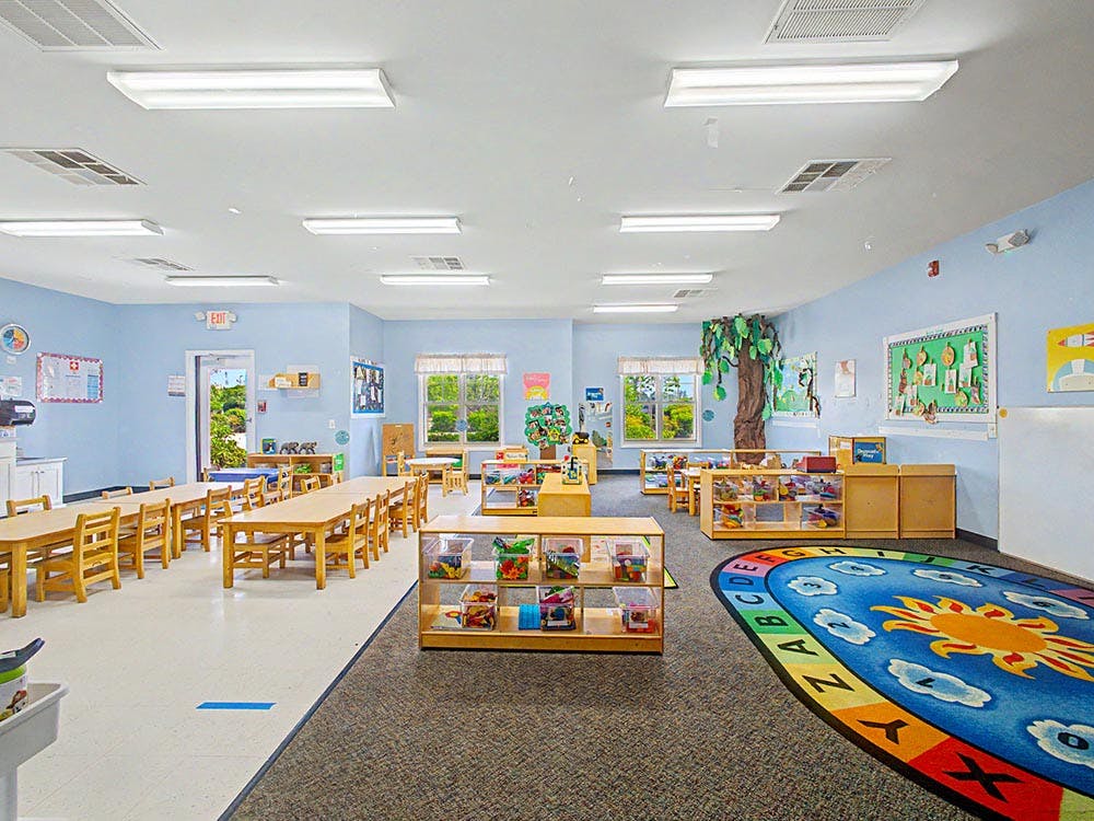 Empty classroom with small tables and chairs, and a colorful rug with ABCs