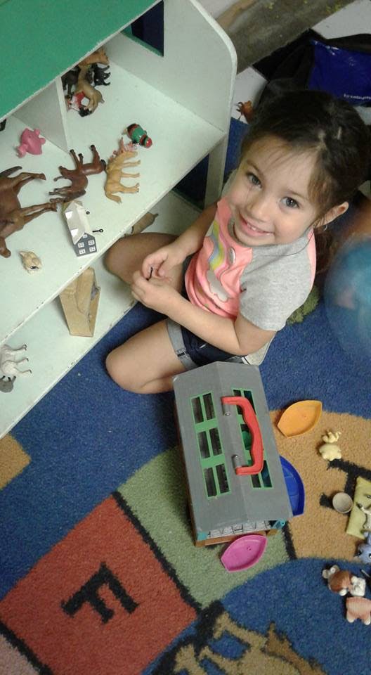 Child sitting on a colorful rug next to a toy shelf and a toy house