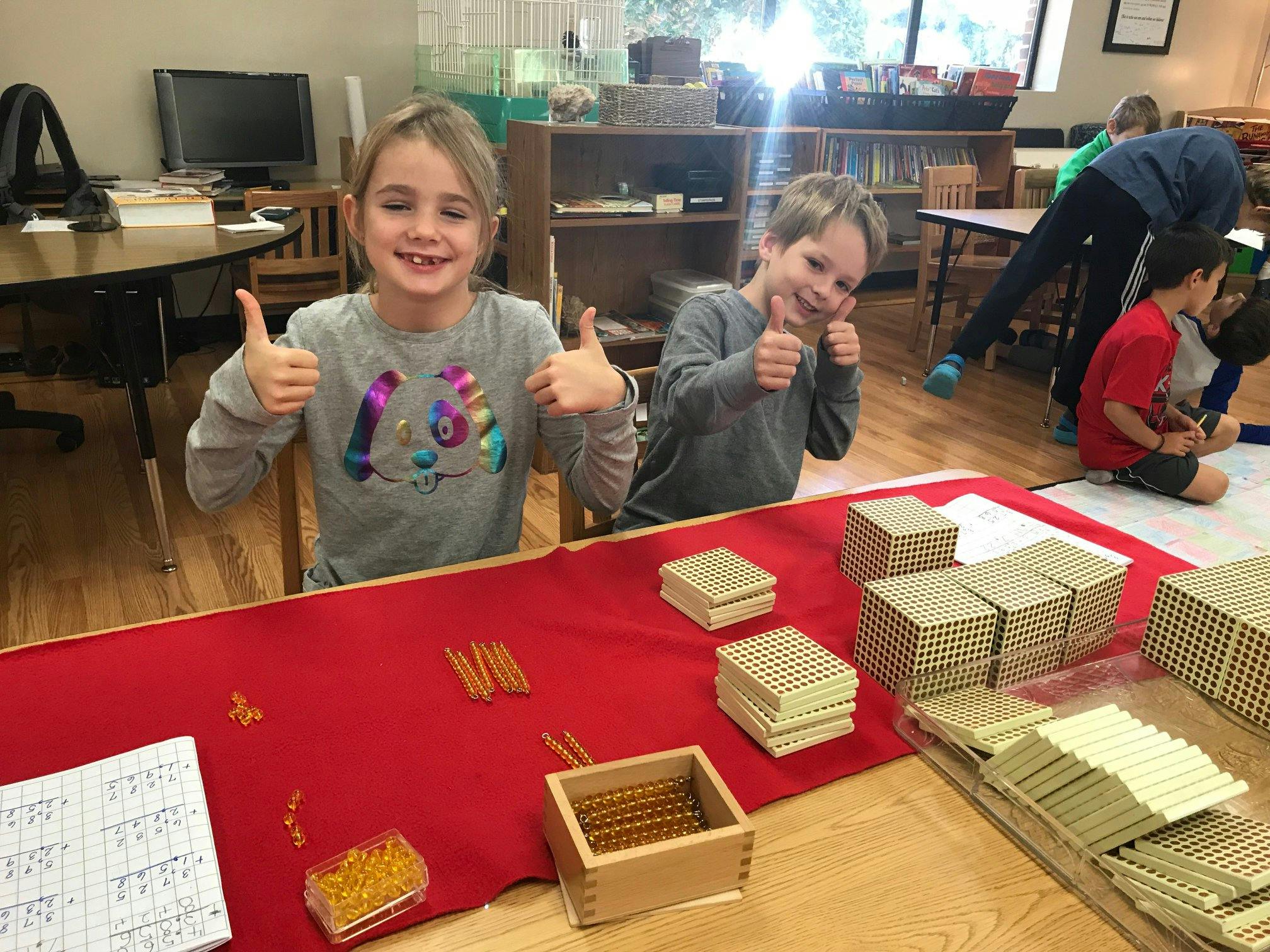 Two children giving thumbs up in a classroom with math manipulatives