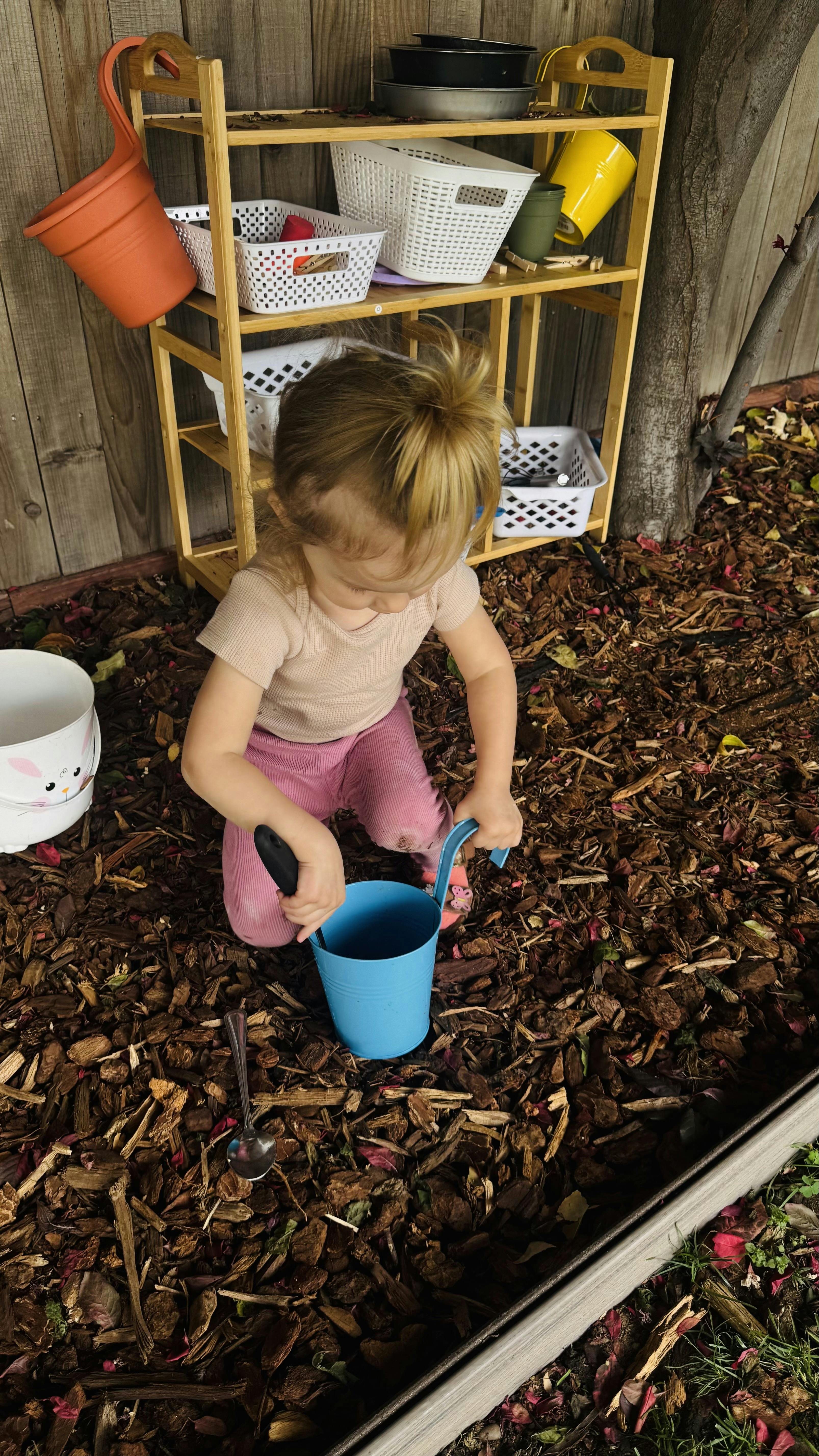 Child kneeling on wood chips, holding a spade in a blue bucket