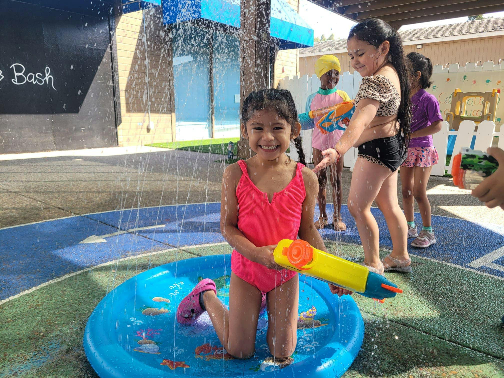 Child in a pink swimsuit kneeling in a small pool with a water gun
