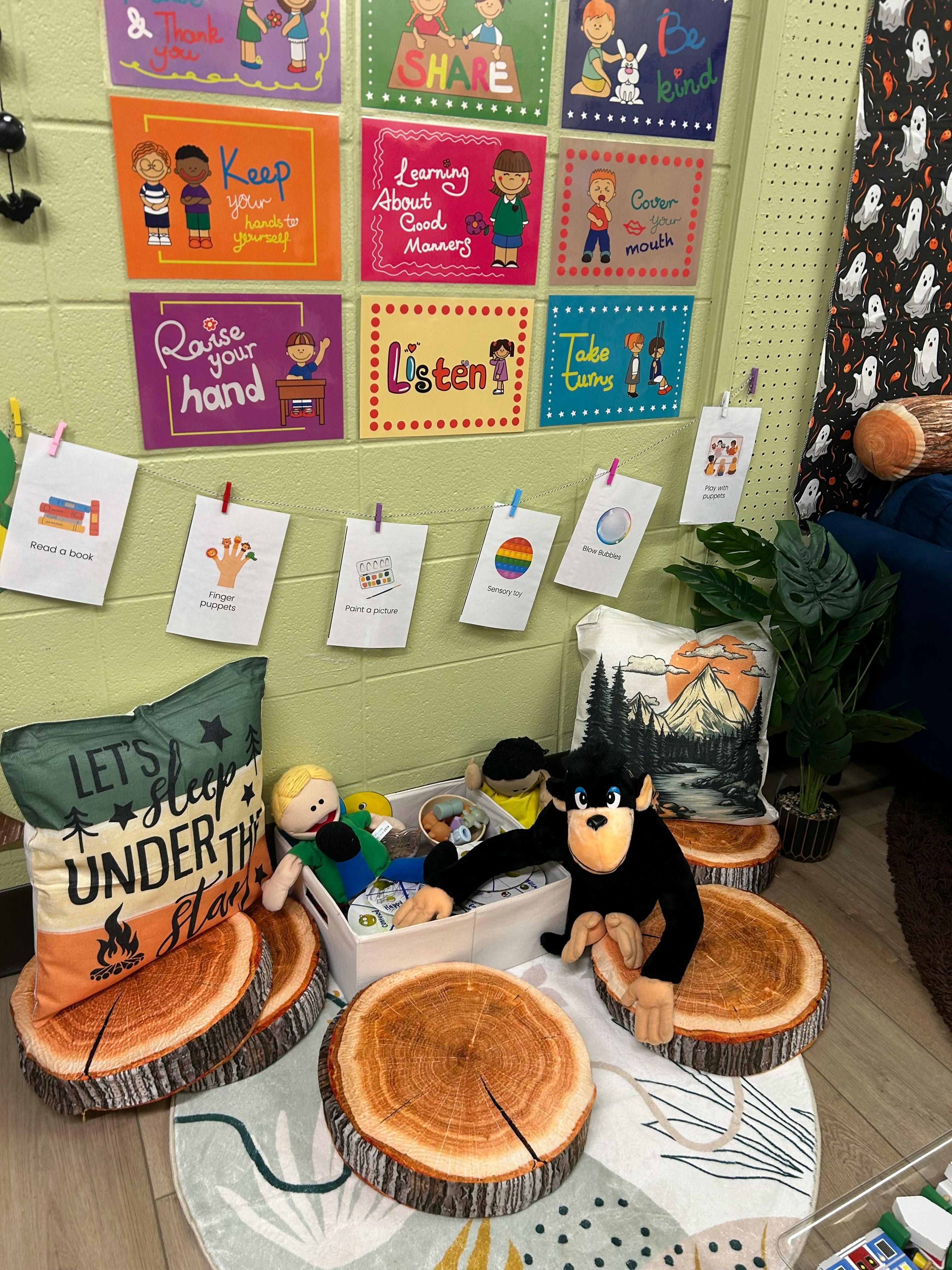 Classroom corner with posters, hanging cards, pillows, stuffed animals, and tree stump seats