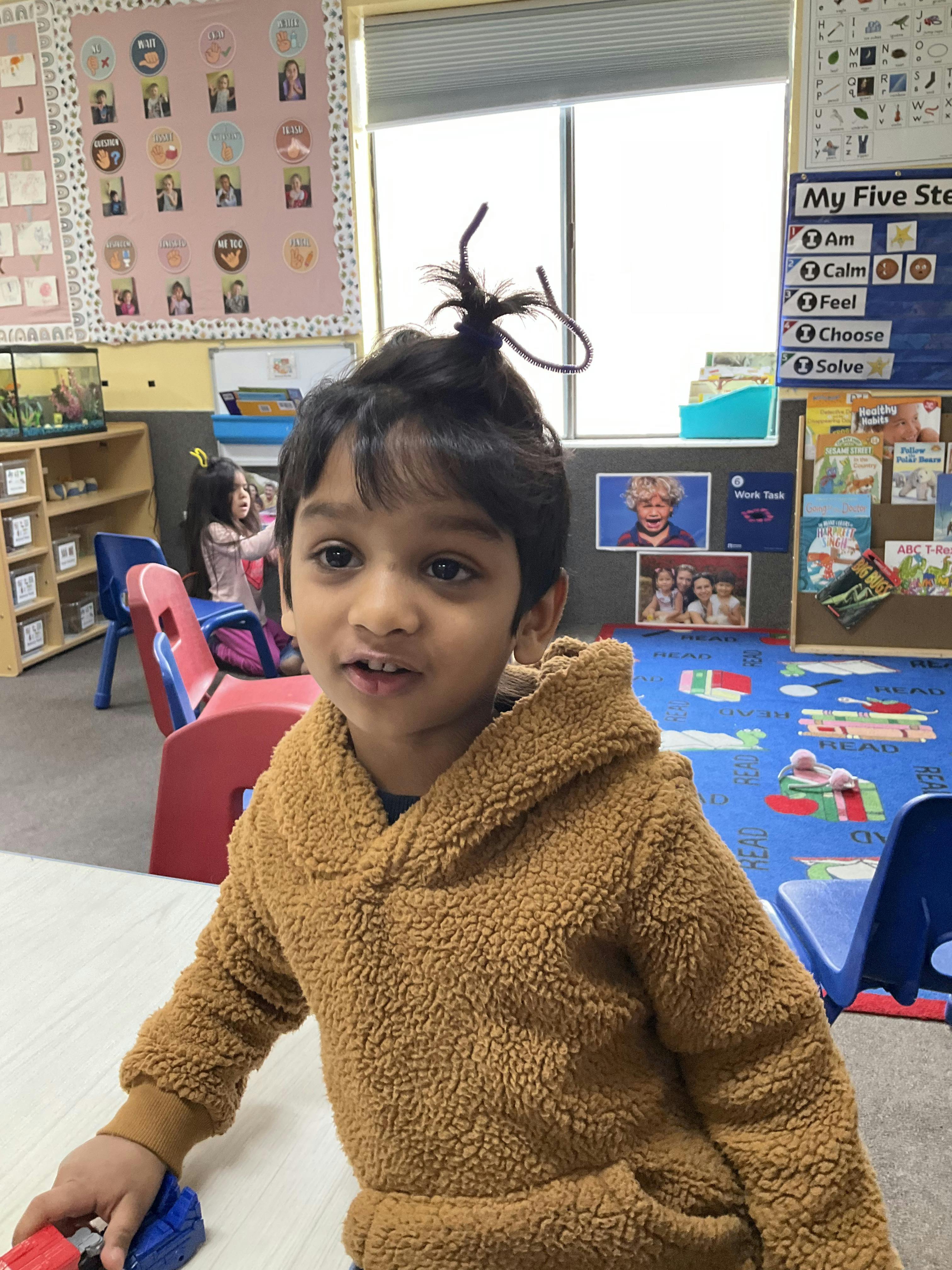 Child in a brown fuzzy hoodie with hair tied up, in a classroom