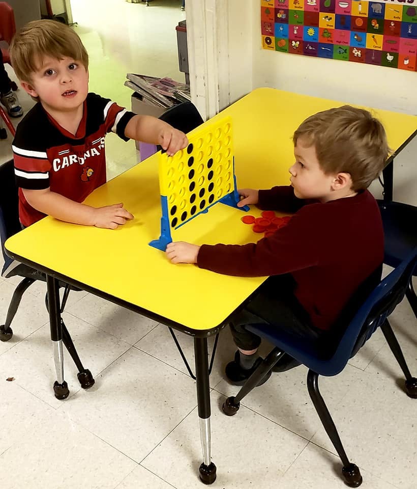 Two children playing a board game at a yellow table in a classroom