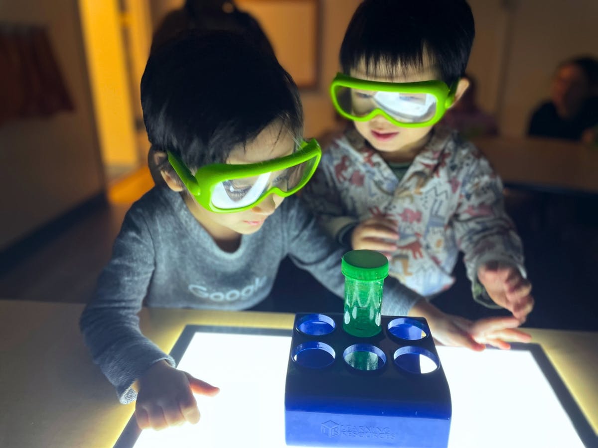 Two children wearing green safety goggles looking at a light table