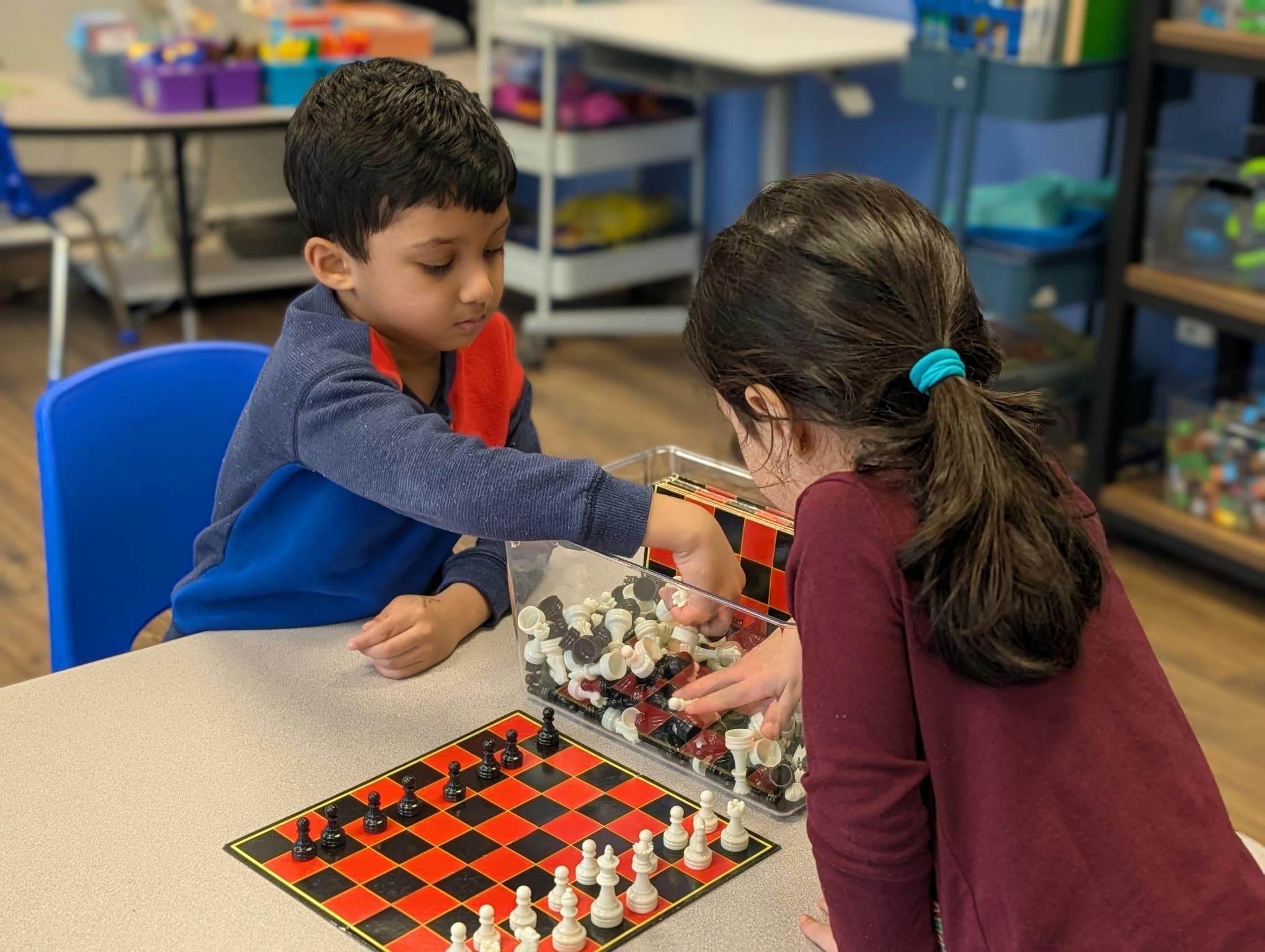 Two children playing with chess pieces and a chessboard in a classroom
