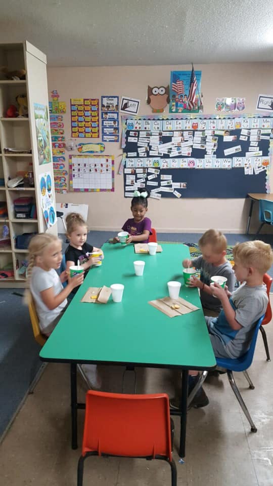 Five children sitting at a green table in a classroom