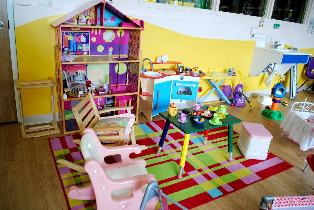 Playroom with dollhouse, toy kitchen, small table with tea set, and chairs