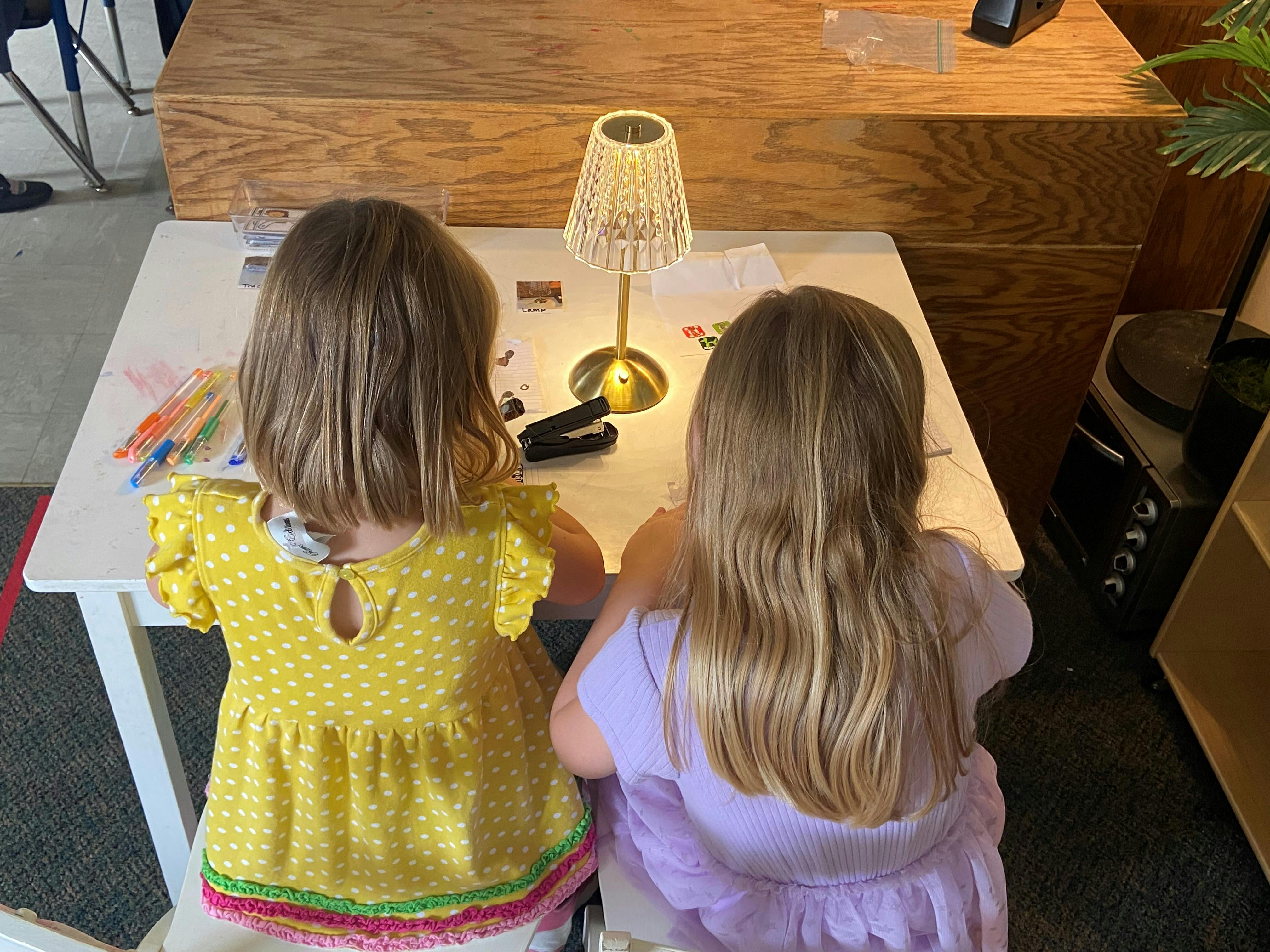 Two children seated at a table with a lamp and art supplies