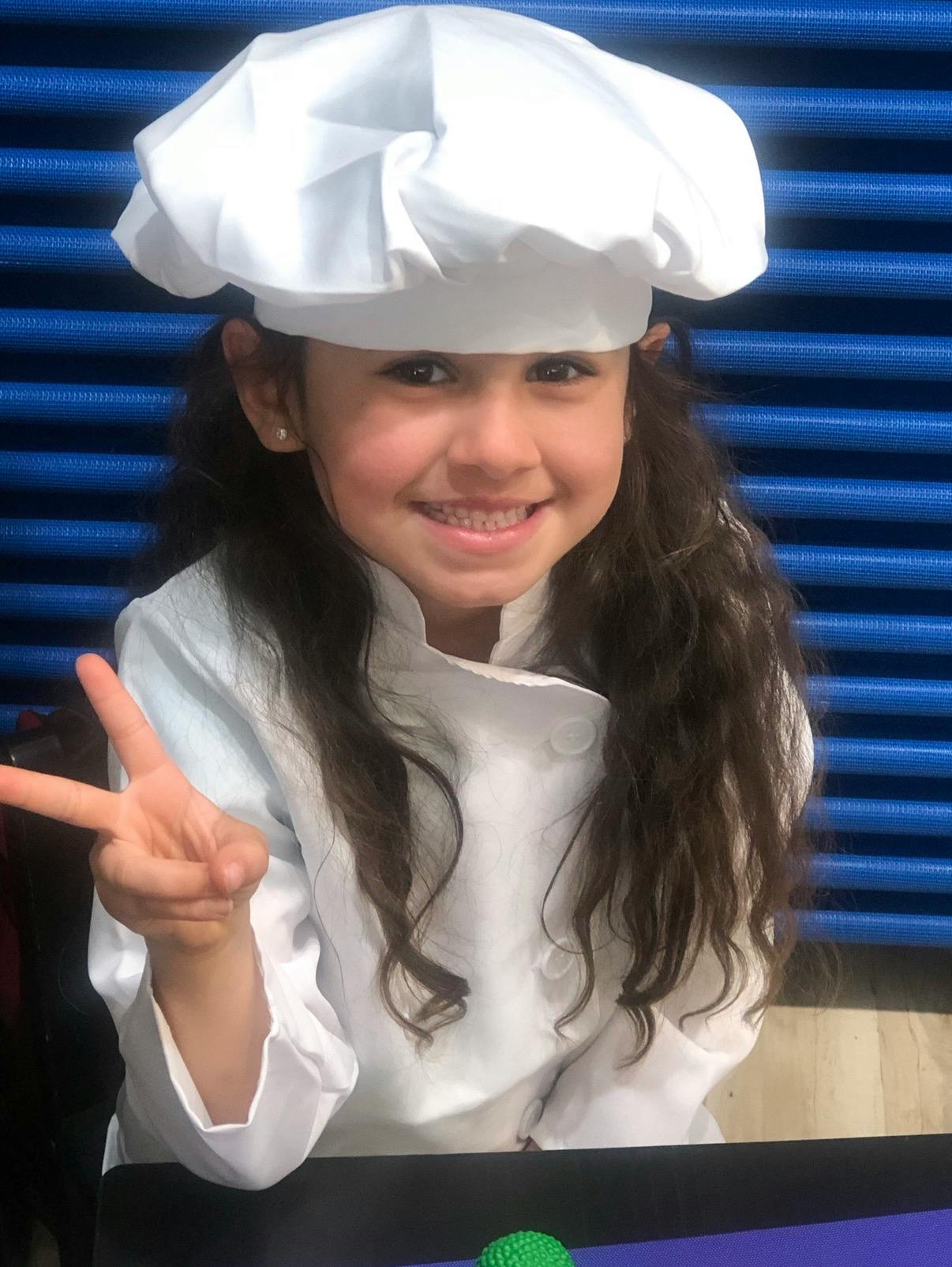Child wearing a white chef hat and jacket making a peace sign