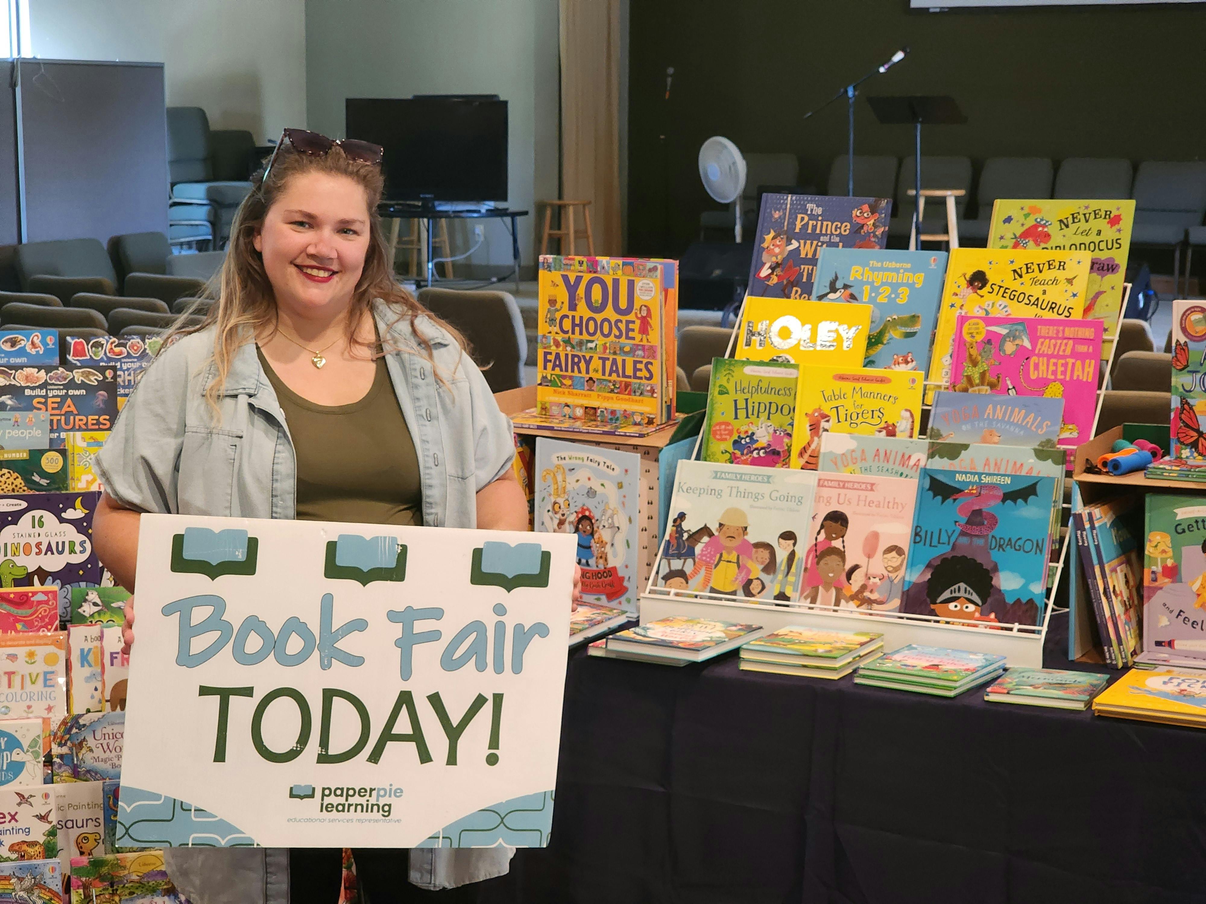 Person standing behind a "Book Fair TODAY!" sign with books displayed