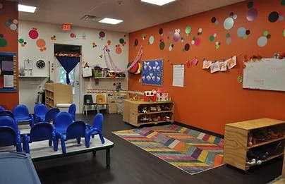 Empty classroom with small blue chairs and tables, colorful rug, and decorated walls