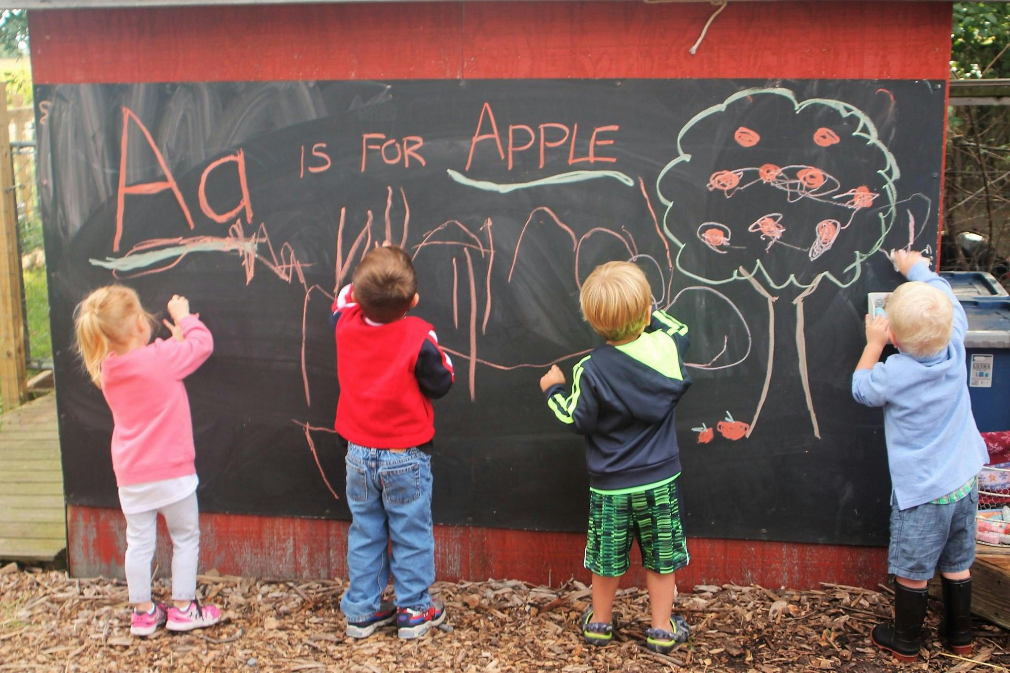 Four children drawing on a large outdoor chalkboard with "A is for Apple" written on it