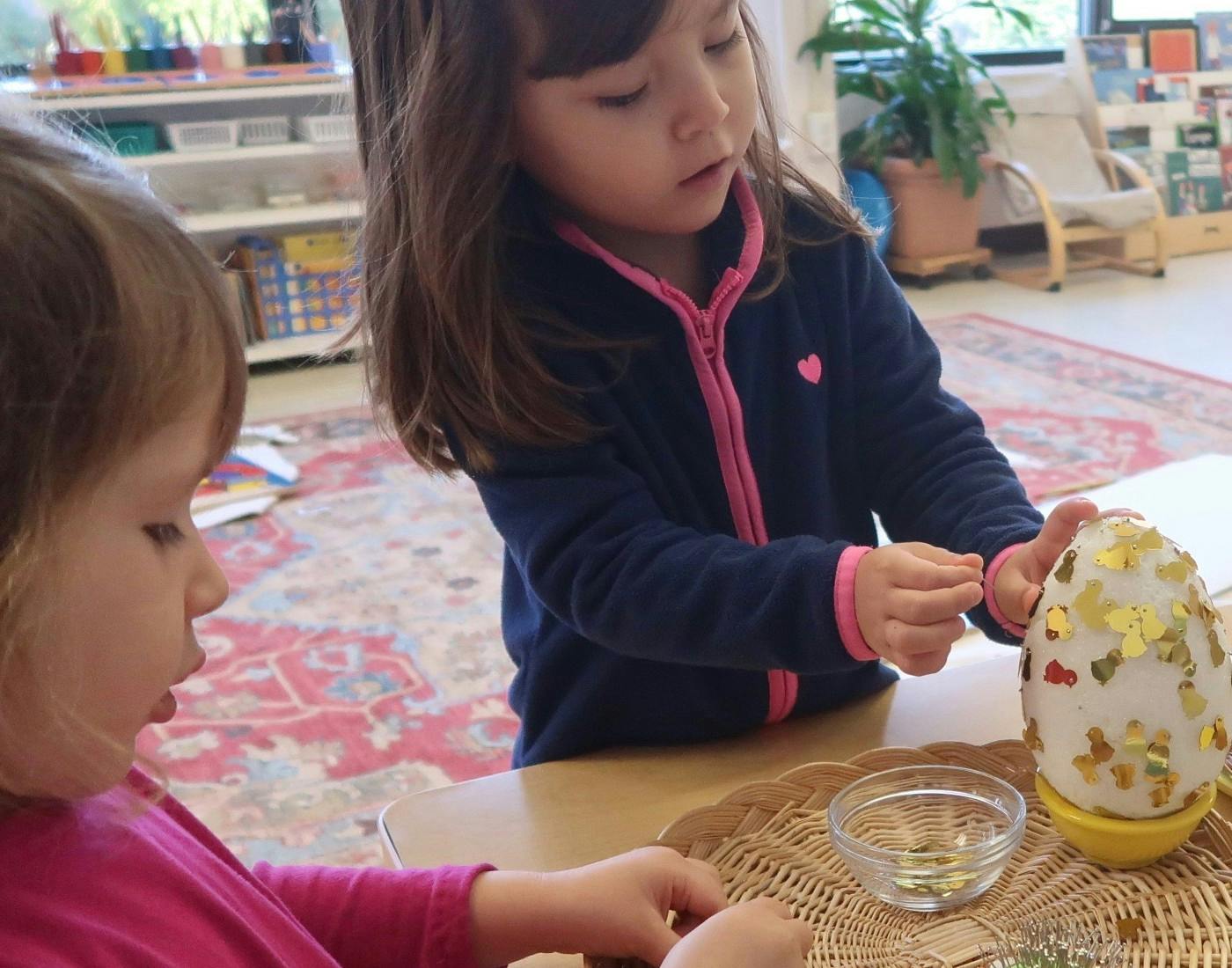 Child decorating an egg with glitter while another child watches