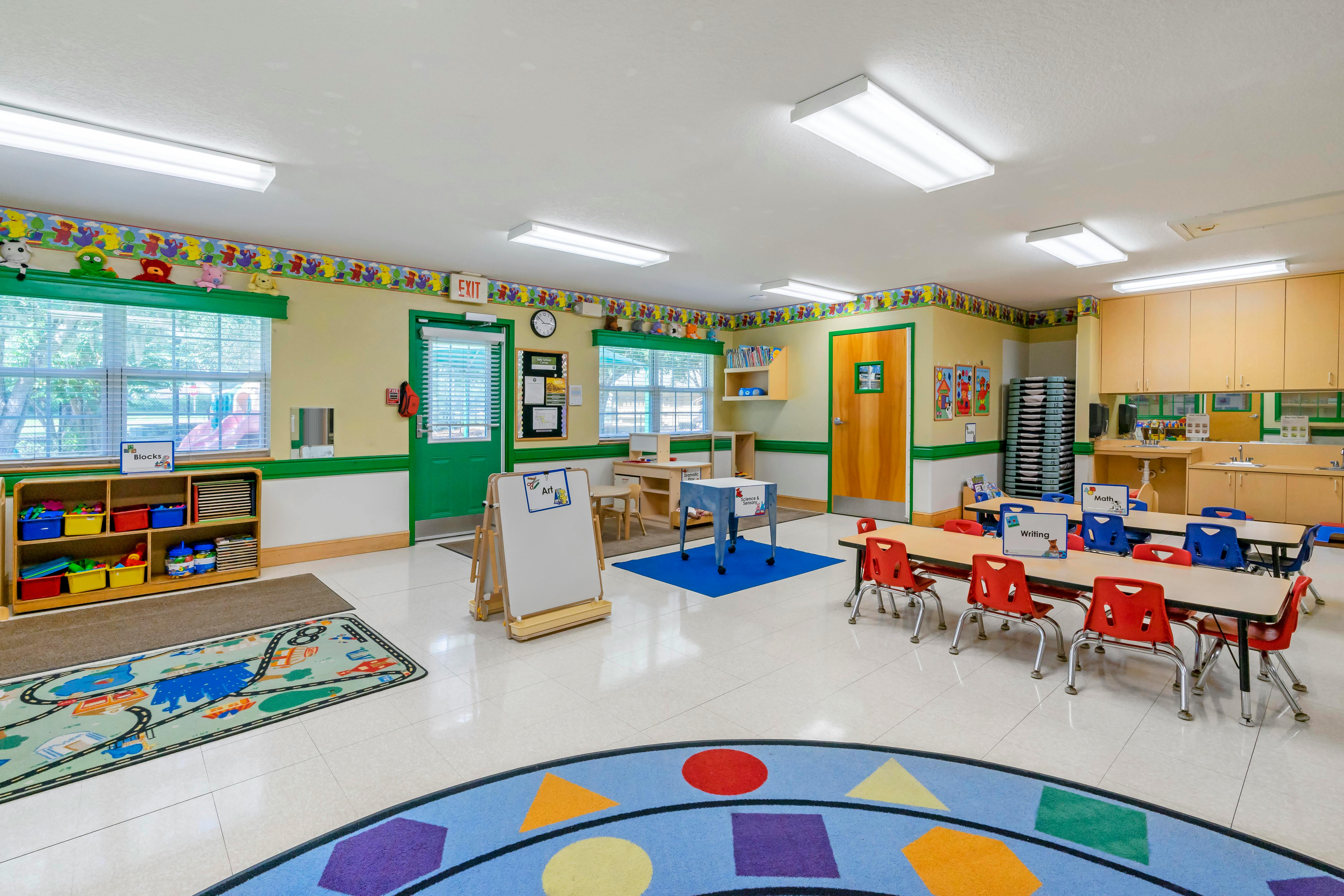 A brightly lit classroom with small tables and chairs, play mats, and learning stations
