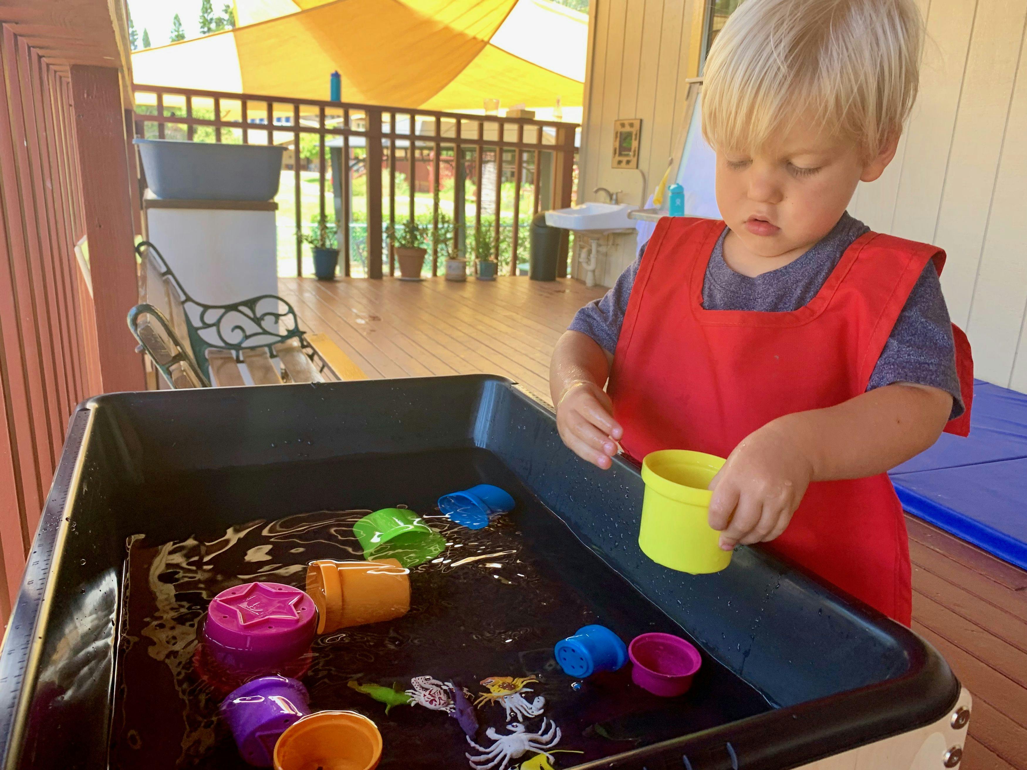 Child in a red apron playing with a yellow cup in a sensory bin