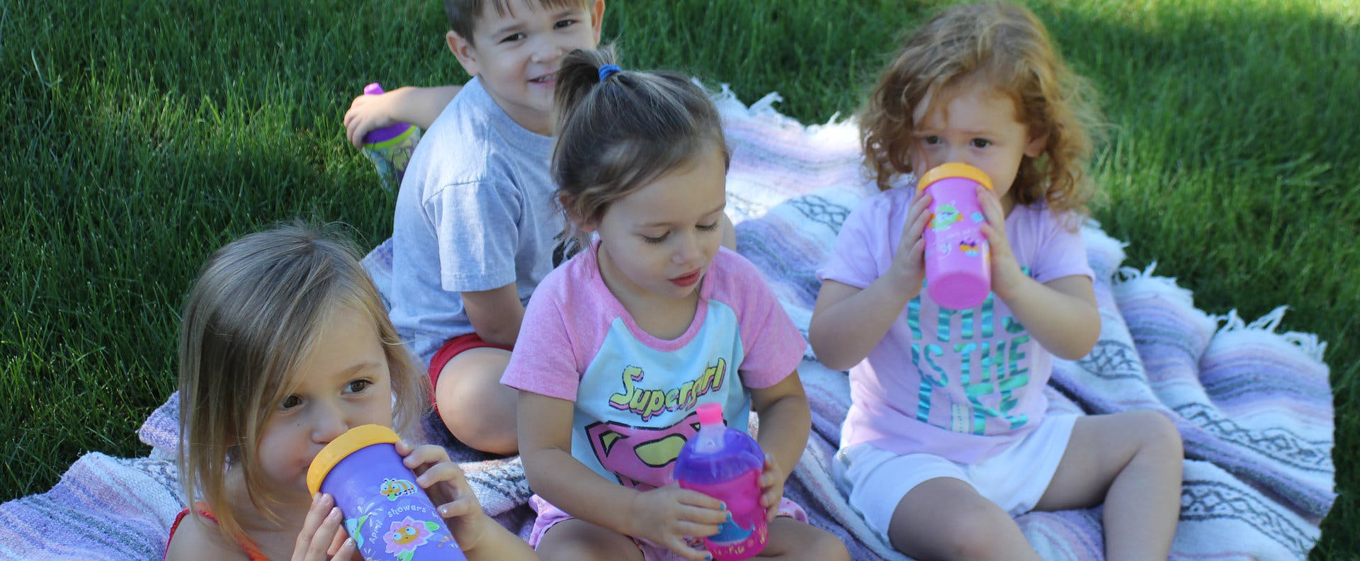 Four children sitting on a blanket, three drinking from sippy cups
