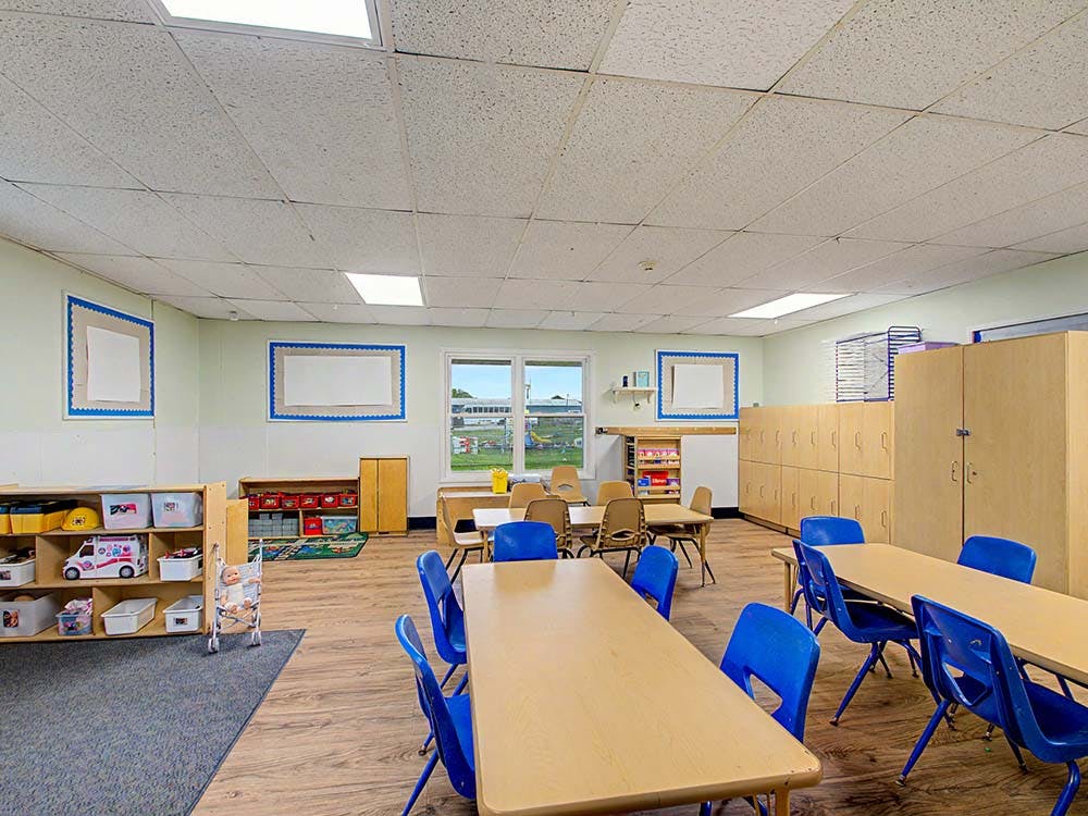 Empty classroom with small tables, blue chairs, cubbies, and toys