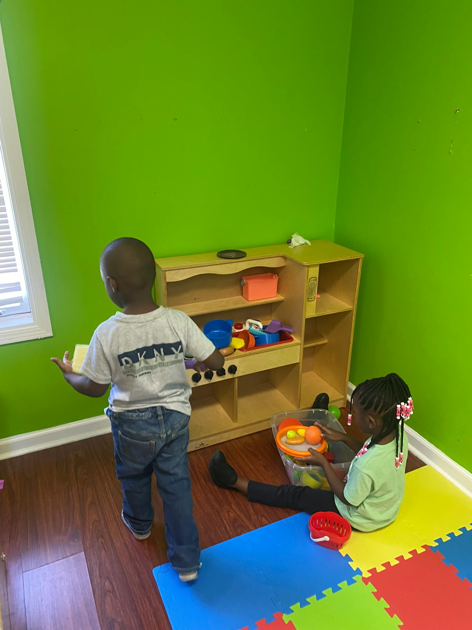Child standing at a play kitchen, another child sitting on the floor playing