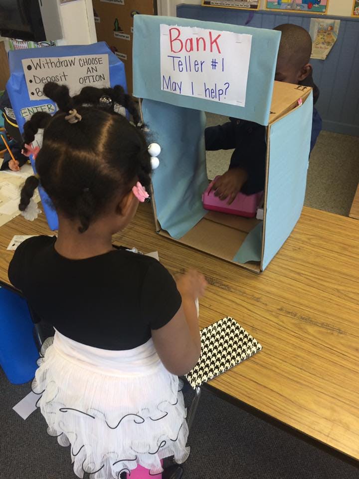 Child at a play bank with a sign that reads "Bank Teller #1 May I help?"