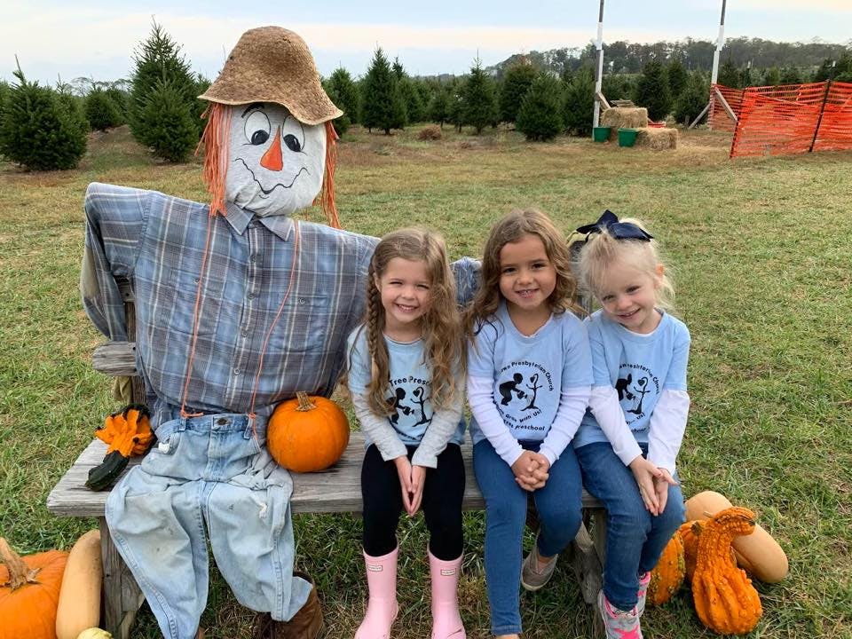 Three children sitting on a bench next to a scarecrow and pumpkins