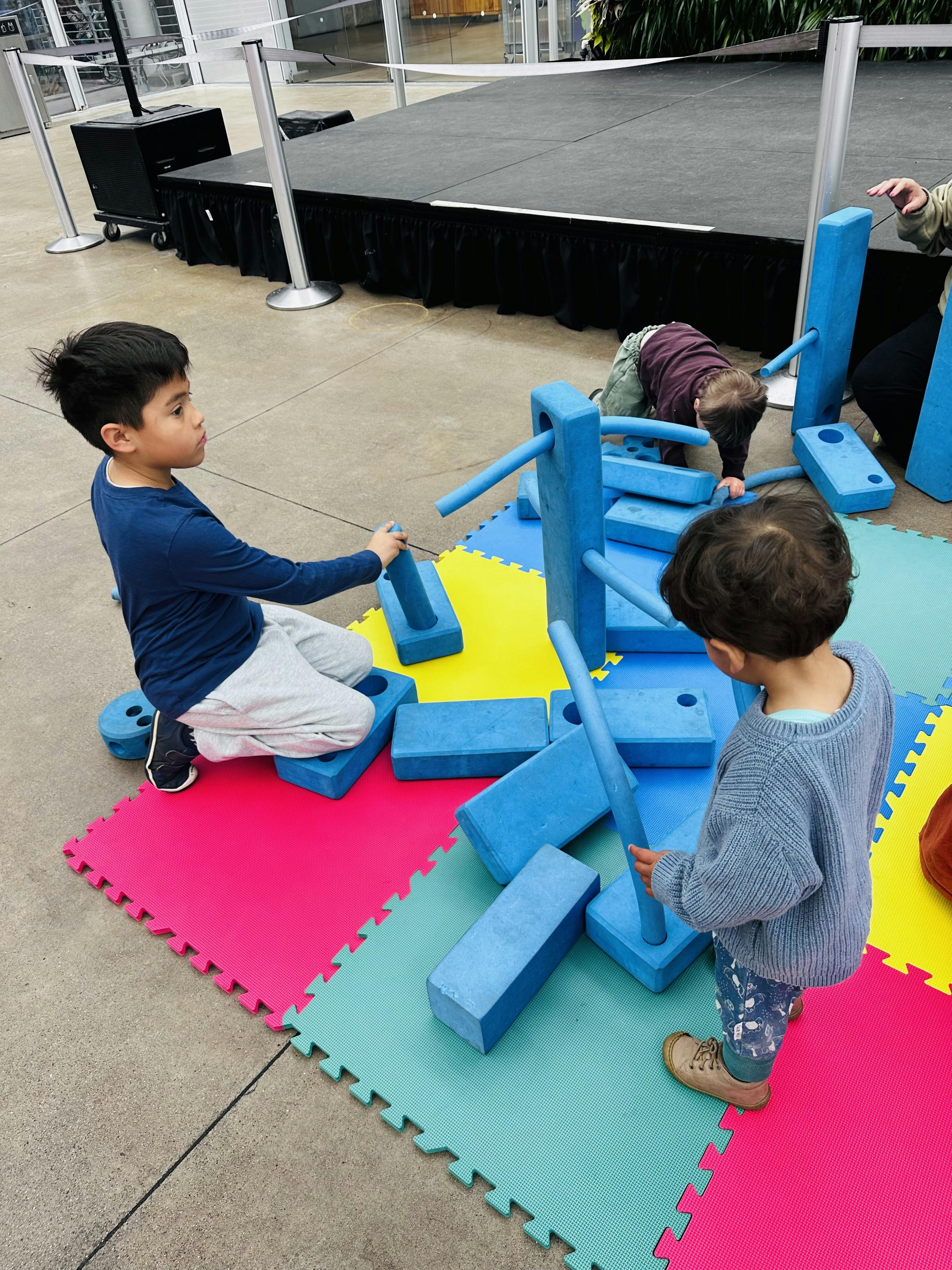 Three children playing with large blue foam blocks on colorful mats