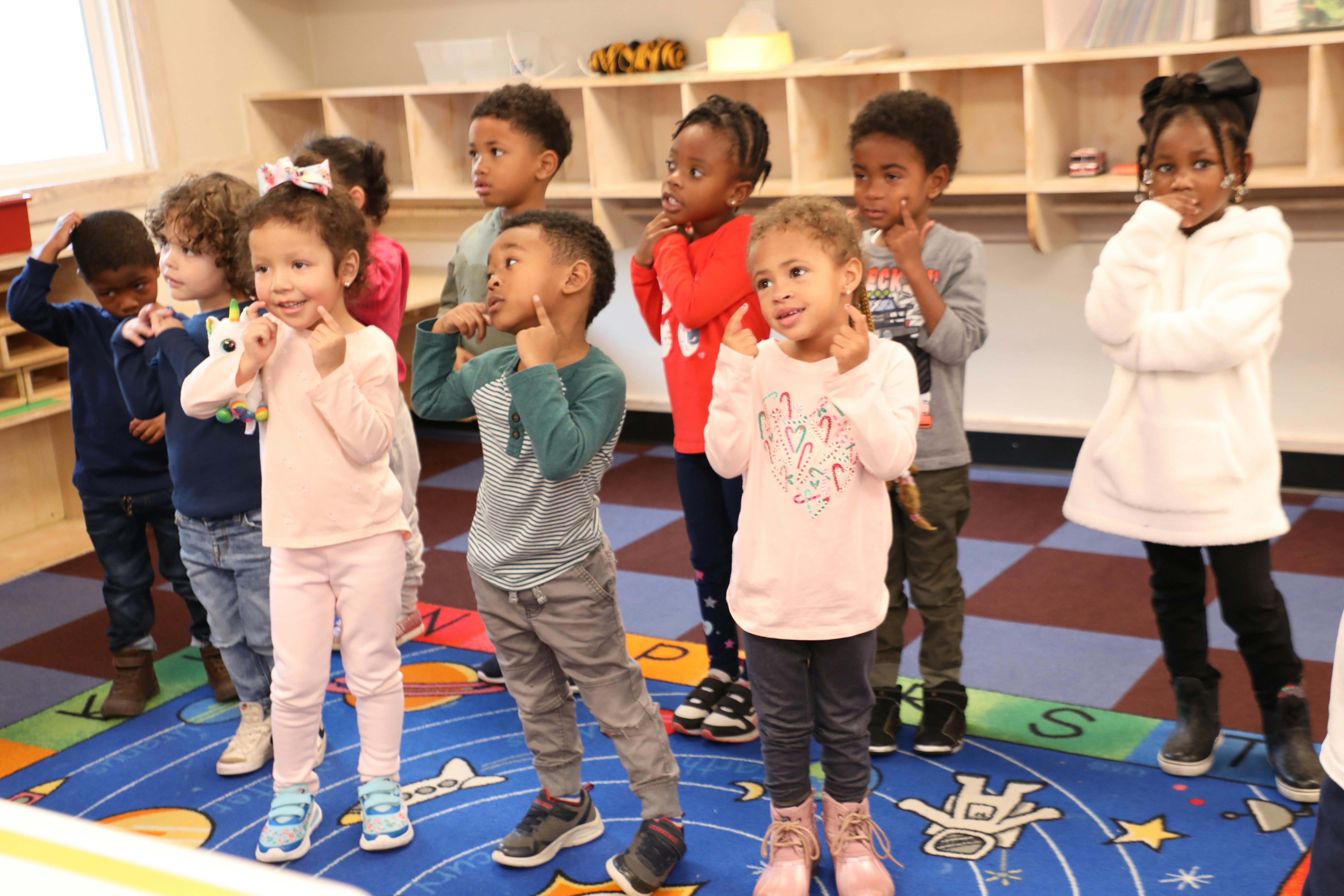 Group of children standing on a colorful rug, some with hands near their faces