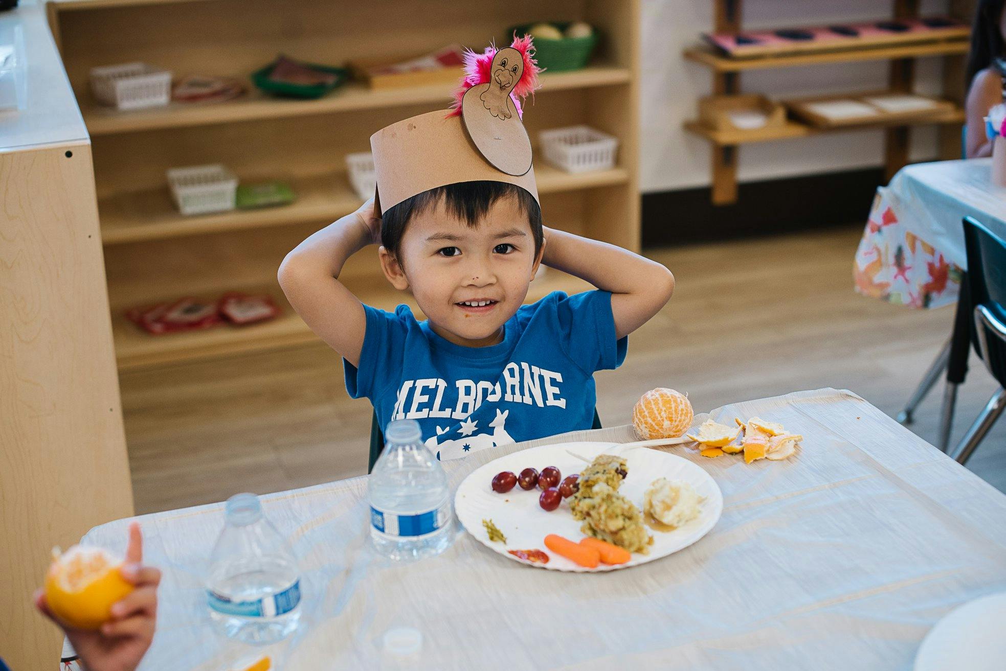 Child wearing a paper hat sitting at a table with a plate of food