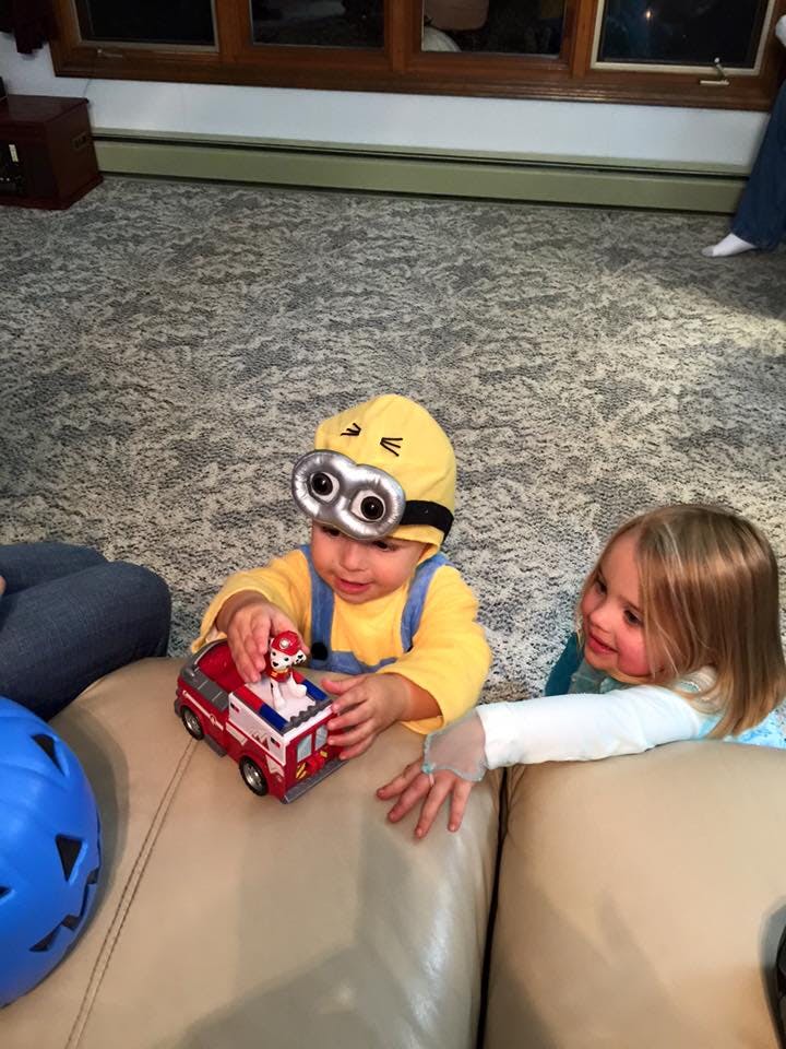 Child in Minion costume playing with a toy fire truck, another child nearby
